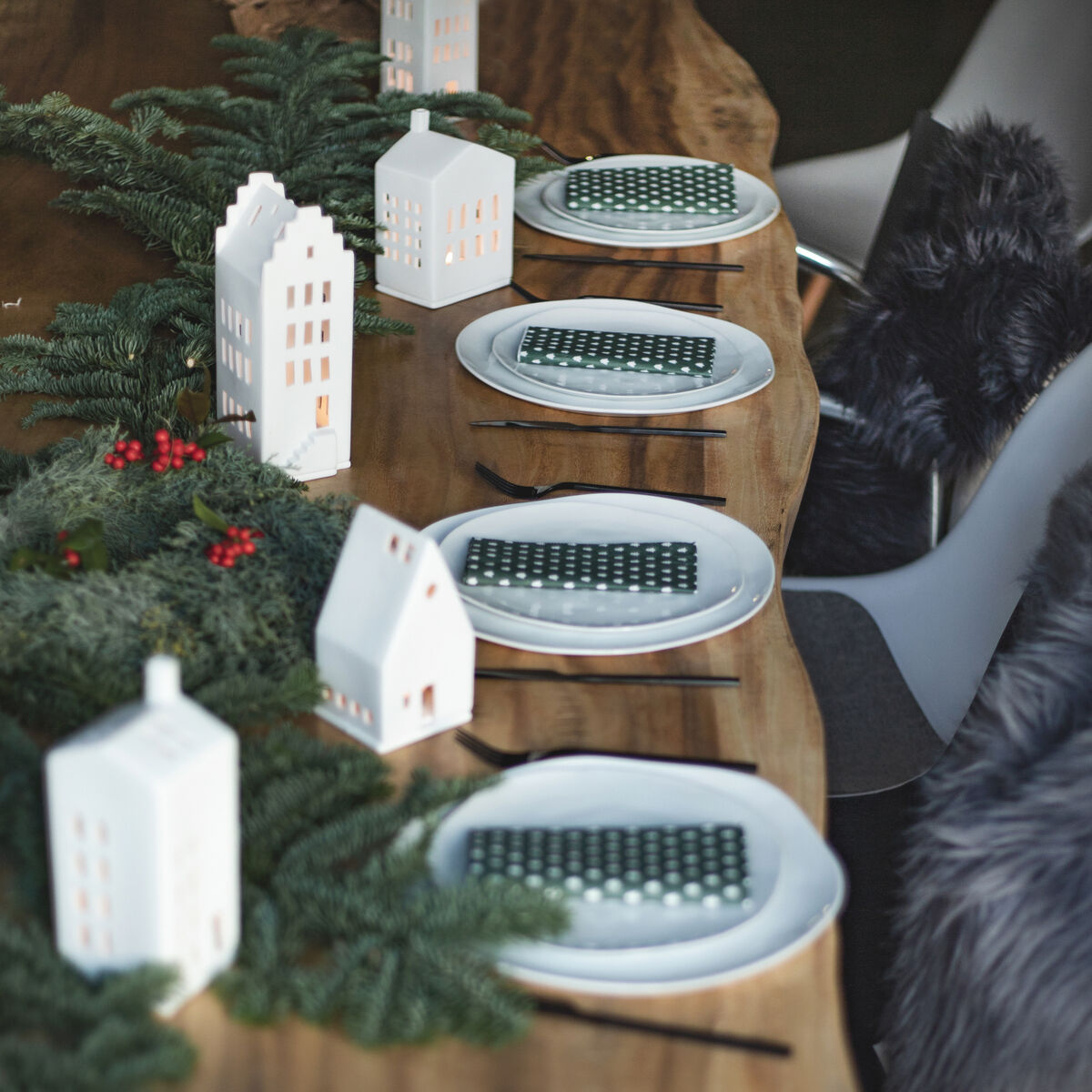 Eine festlich gedeckte Tischdekoration mit weißen Keramik-Teelichthäusern in Form von Giebeldachhäusern, umgeben von grünen Tannenzweigen und roten Beeren. Der Tisch ist aus Holz mit einer rustikalen Oberfläche und gedeckt mit weißem Geschirr und grün-weiß gepunkteten Servietten.