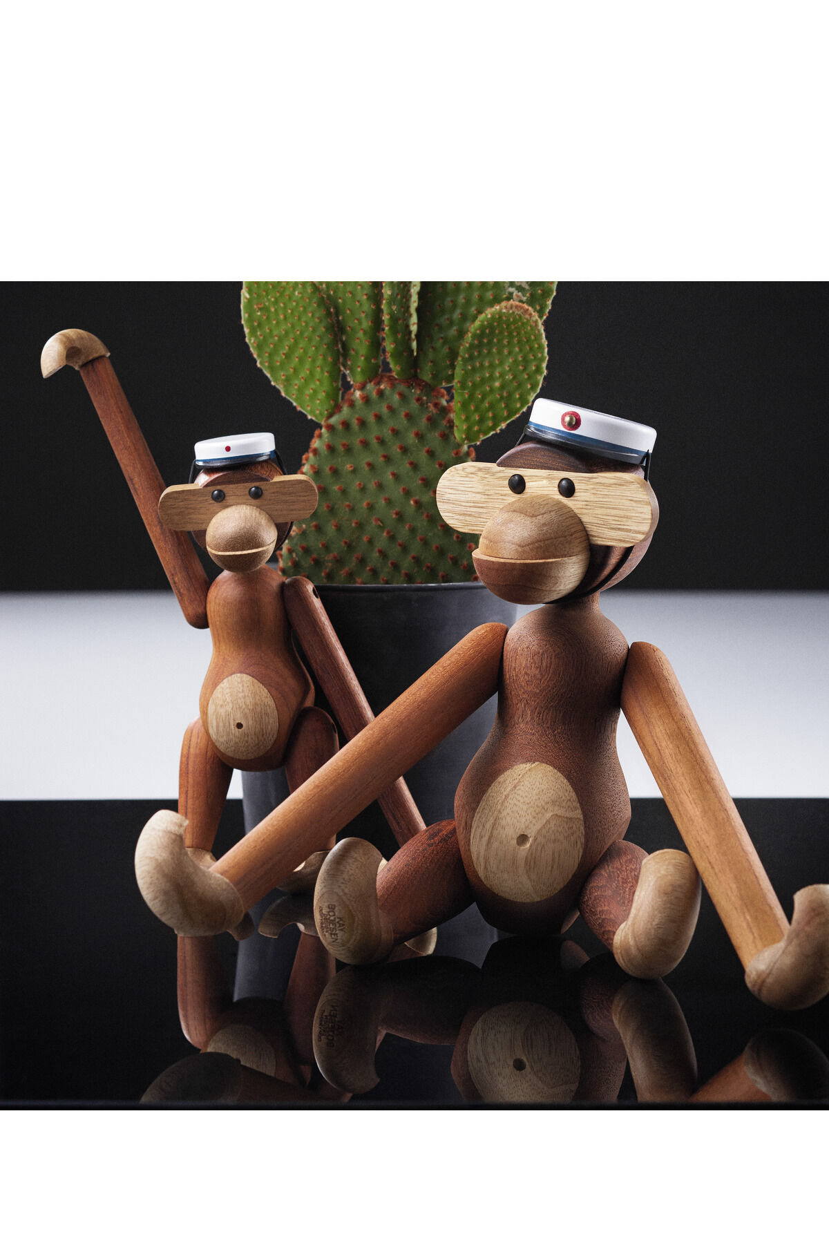 Zwei Holzfiguren in Form von Affen von Kay Bojesen, eine stehend und eine sitzend, vor einem Kaktus auf einem schwarzen Hintergrund. Die Figuren tragen Mützen mit weißem und schwarzem Band und rotem Knopf.