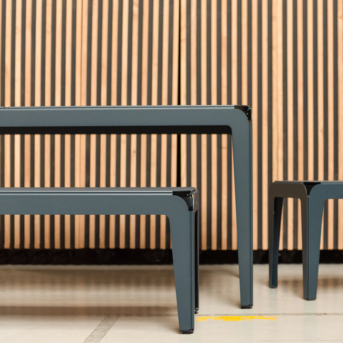 Ein modernes Gartentisch-Set namens Bended Table, bestehend aus einem großen und einem kleineren Tisch, mit einem minimalistischen, kantigen Design aus Metall, aufgestellt vor einer hölzernen Wand in warmen Brauntönen.