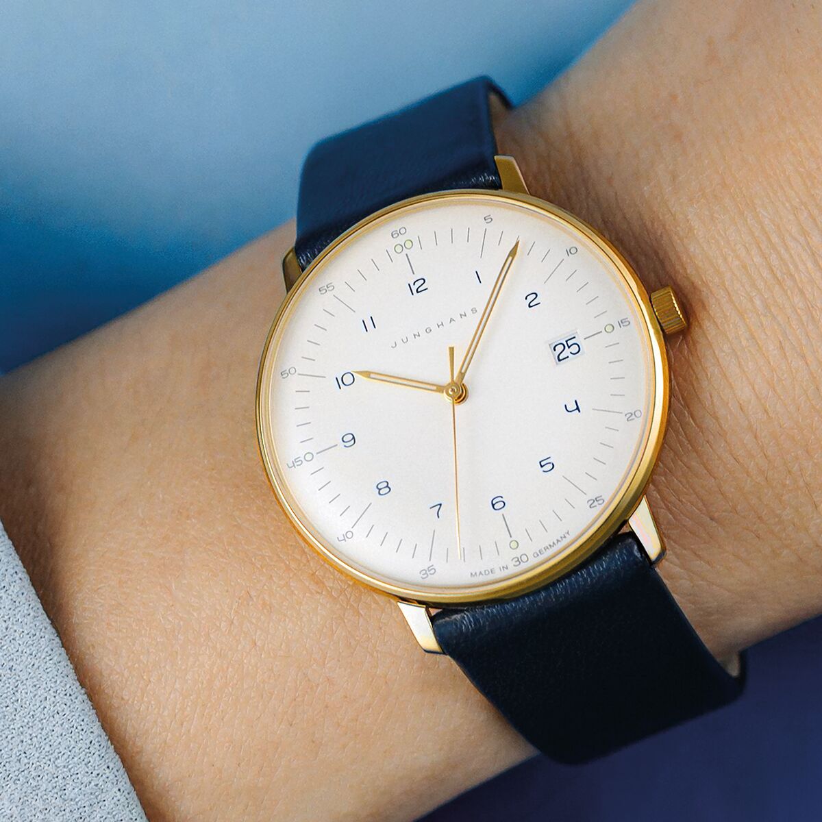 Elegante Damenarmbanduhr der ZEIT Sonderedition »max bill Damen« von Junghans mit weißem Zifferblatt, schlanken goldfarbenen Indizes sowie einem Gehäuse aus Edelstahl in Softgold und einem dunkelblauen Lederarmband. Die Uhr wirkt minimalistisch und ist auf 100 Stück limitiert.