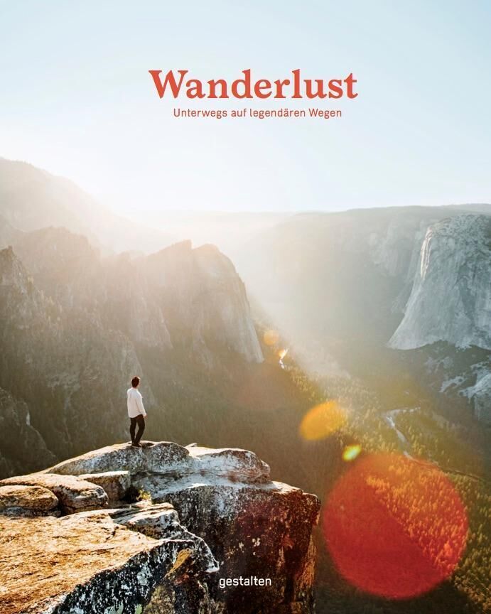 Eine Person steht auf einem hohen Felsen und blickt in eine weite, bergige Landschaft im Sonnenaufgang. Der Titel des Buches 'Wanderlust' ist oben zu sehen.