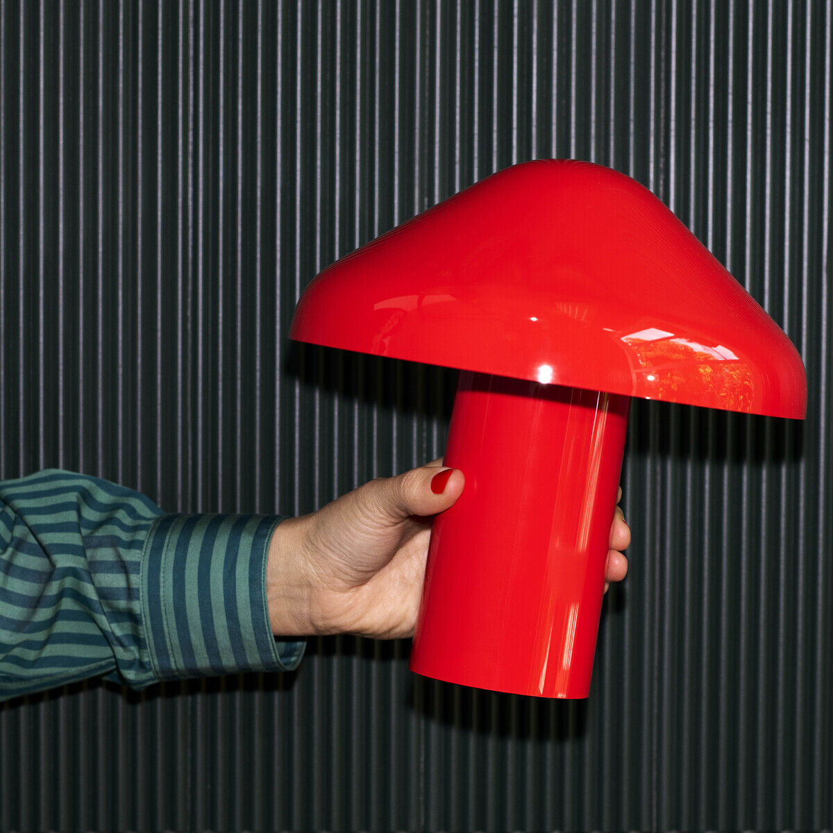 Eine tragbare Lampe mit modernem Design in Form eines Pilzes. Die Lampe hat eine leuchtend rote Farbe und wird von einer Hand gehalten. Der Hintergrund ist dunkel und geriffelt.