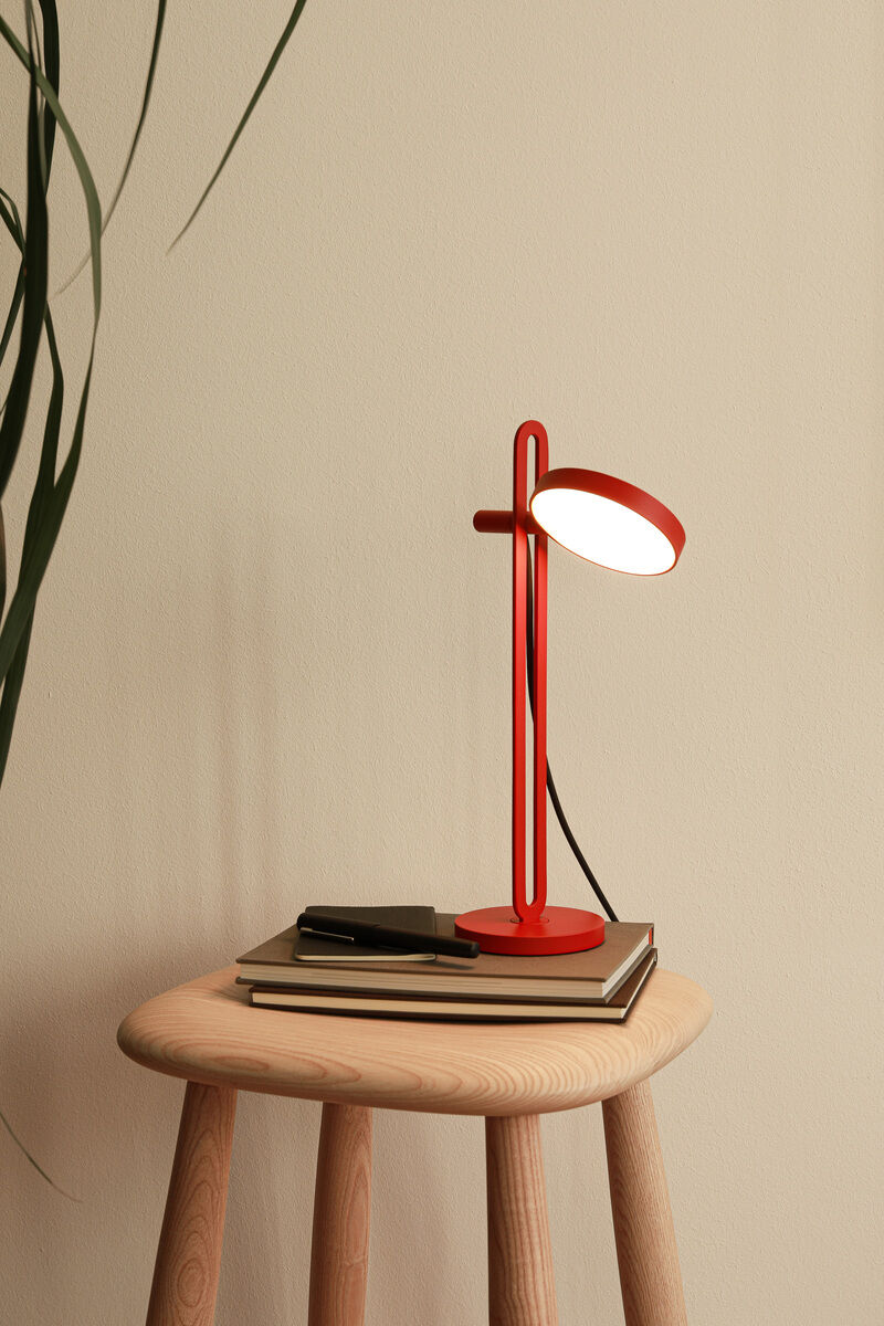Eine moderne, rote Tischlampe namens »Echo« steht auf einem runden Holztisch. Die Lampe hat ein minimalistisches Design und ein LED-Licht, das den Tisch darunter beleuchtet. Neben der Lampe liegen Bücher und ein Smartphone auf dem Tisch. Der Hintergrund ist beige mit einem Teil einer Pflanze am linken Rand.