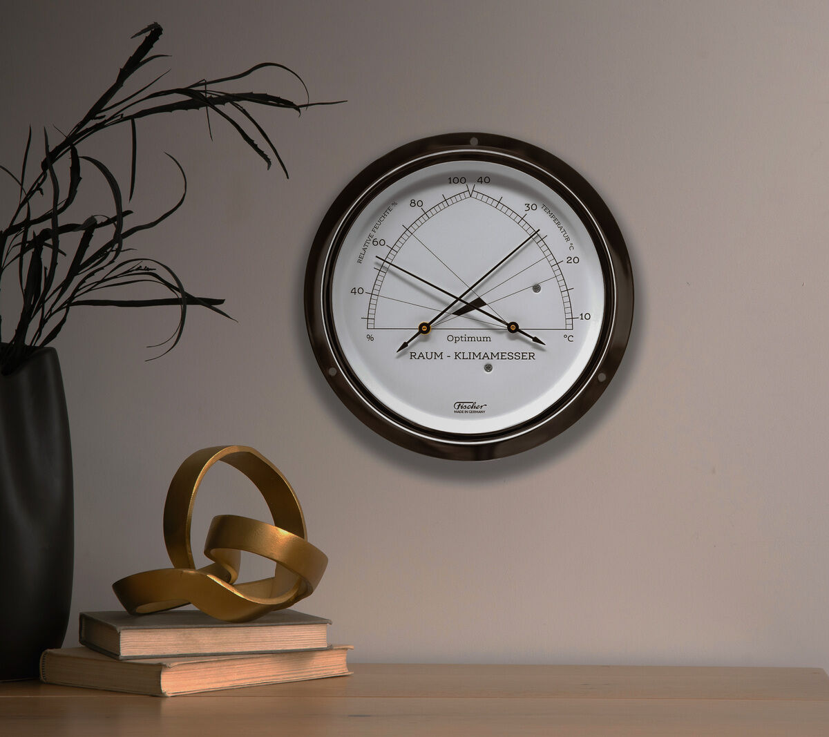 ZEIT-Sonderedition »Raumklimamesser« an einer grauen Wand montiert, neben einer Vase mit schwarzen Zweigen und einem Holztisch mit goldener Dekoration darauf.