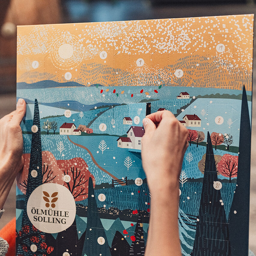 Adventskalender »Winterlandschaft« von Ölmühle Solling mit stimmungsvoller Illustration einer verschneiten Landschaft, Tannenbäumen, Häusern und 24 nummerierten Türchen. Hände öffnen ein Türchen. Farben: Blau-, Weiß- und Goldtöne, detailreich und festlich gestaltet.