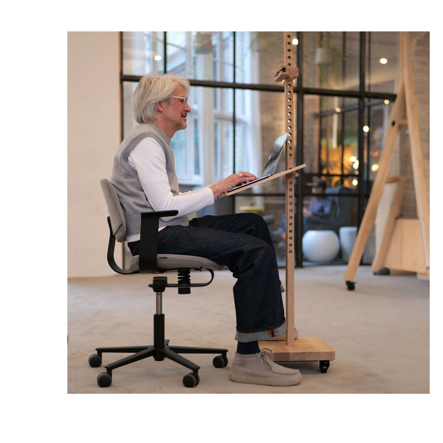 Ein älterer Mann sitzt auf einem ergonomischen Bürostuhl vor einem mobilen Device-Stativ »ERA« aus naturbelassenem Eichenholz. Auf der neigbaren Ablage steht ein Laptop. Die moderne, helle Wohnung hat freiliegende Steinwände, Glasflächen und viel Licht.