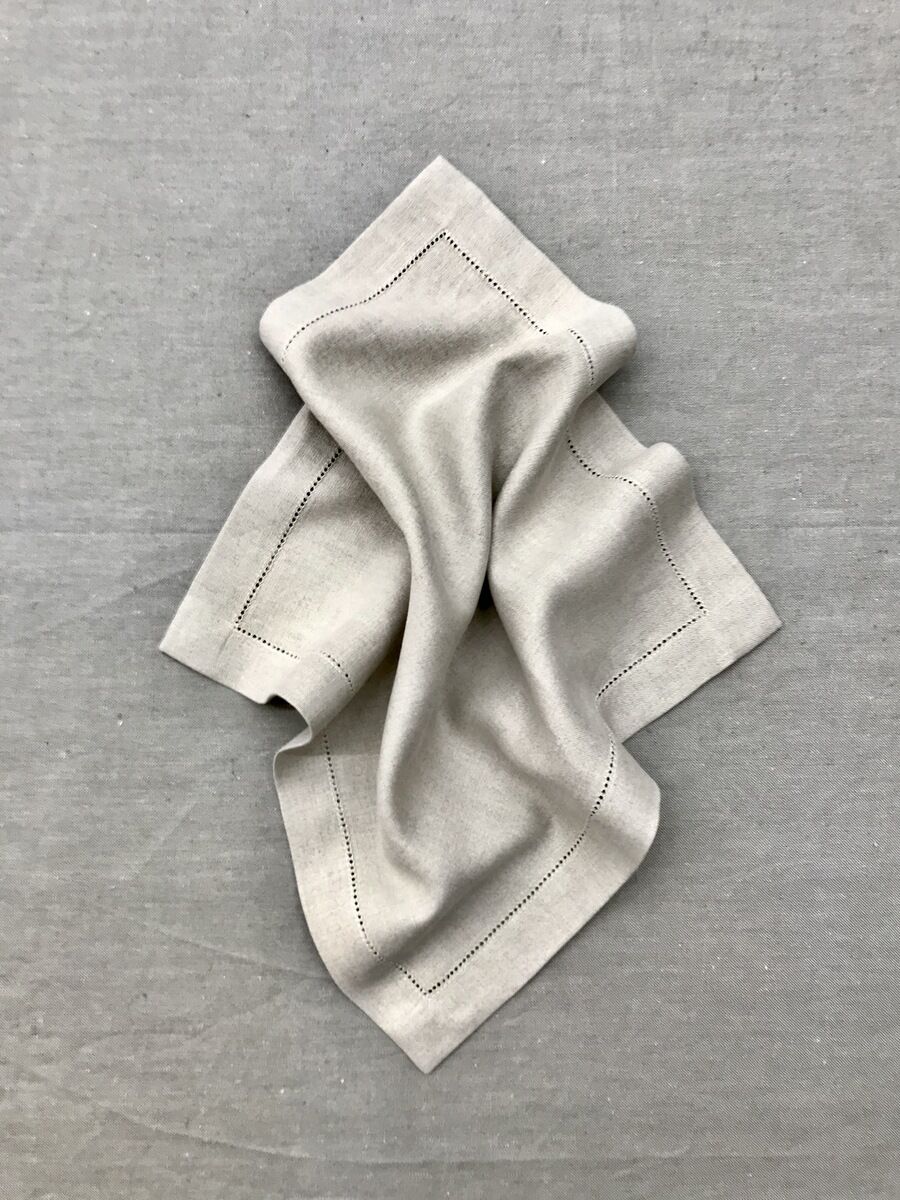 Eine beiger Leinen-Serviette liegt kunstvoll drapiert auf einer gleichfarbigen grauen Oberfläche. Die Serviette ist schlicht und elegant, mit einer feinen, gestickten Einfassung entlang der Ränder.