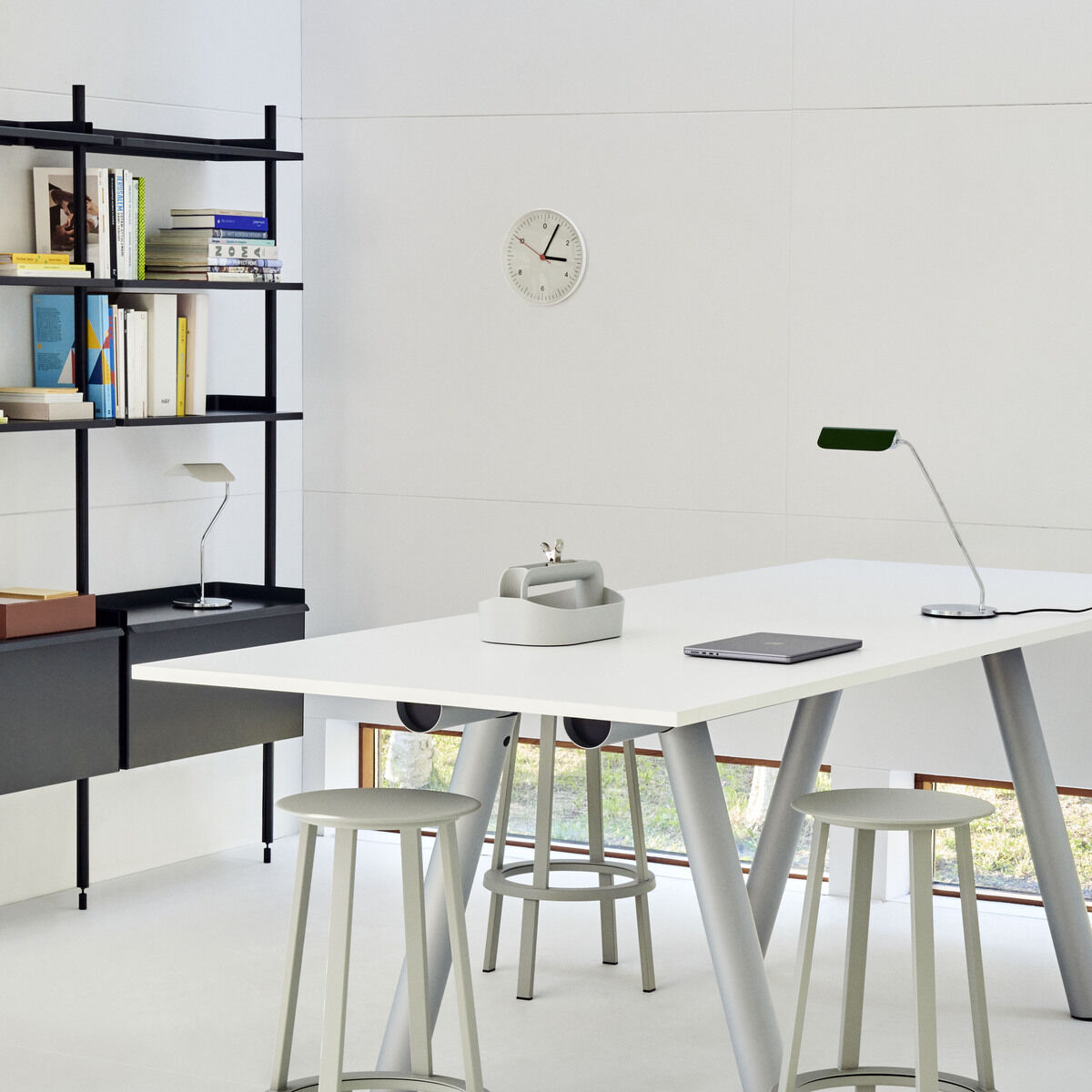 Moderne, minimalistische Arbeitsumgebung mit weißem Schreibtisch und der Apex Lampe »Desk« darauf. Schwarzes Bücherregal im Hintergrund, graue Hocker und gut beleuchteter Raum mit großen Fenstern.