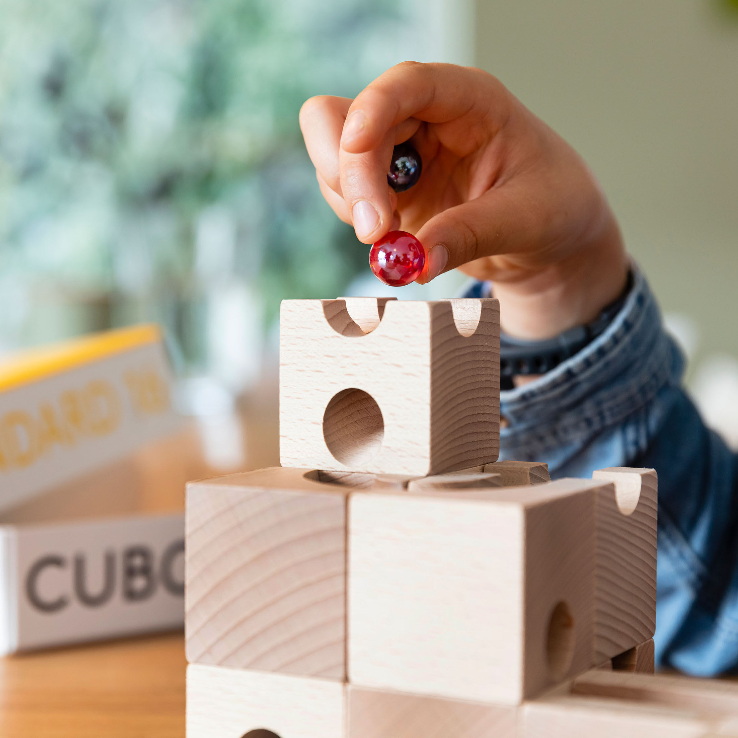 Ein Kind hält zwei bunte Glasmurmeln über einer aufgebauten Kugelbahn aus naturbelassenem Buchenholz der Marke Cuboro. Im Hintergrund sind eine leicht verschwommene Verpackung und der Schriftzug "STANDARD 50" zu erkennen.