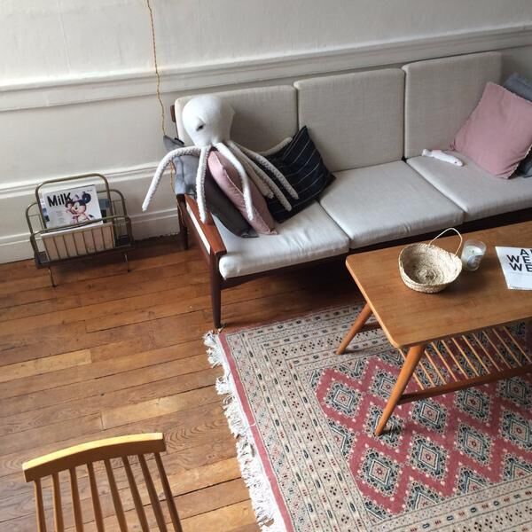 Ein weißes Plüschtier in Form eines Albino-Oktopus liegt auf einem grau-beigen Sofa mit rosa und schwarzen Kissen. Das Wohnzimmer ist mit einem Holzboden und einem Teppich dekoriert. Ein flacher Holztisch steht vor dem Sofa.