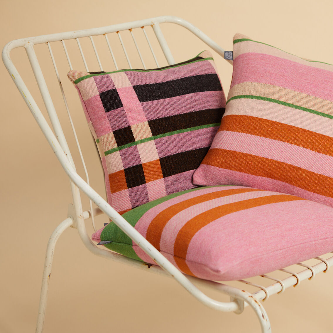 Das Bild zeigt drei farbenfrohe Kissen auf einem weißen Metallstuhl. Die Kissen haben geometrische Muster und sind in Rosa-, Orange-, Grün- und Schwarztönen gehalten.