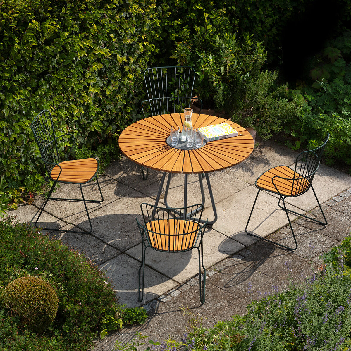 Ein Gartentisch namens CIRCLE mit einem runden Holz-Tischblatt und vier passenden Stühlen mit Metallgestellen steht auf einer gepflasterten Terrasse, umgeben von üppigem Grün und Hecken.