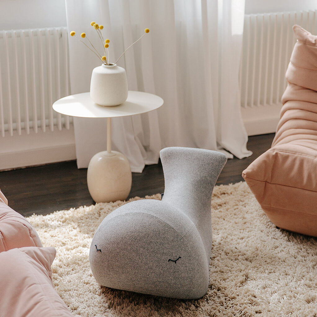 Ein modern gestalteter Schaukelwal in grauer Farbe steht auf einem beigen, flauschigen Teppich. Im Hintergrund sind ein weißer, runder Tisch mit einer weißen Vase und gelben Blumen sowie rosa Sitzkissen zu sehen. Die Szene befindet sich in einem hellen Raum mit einem großen Fenster.