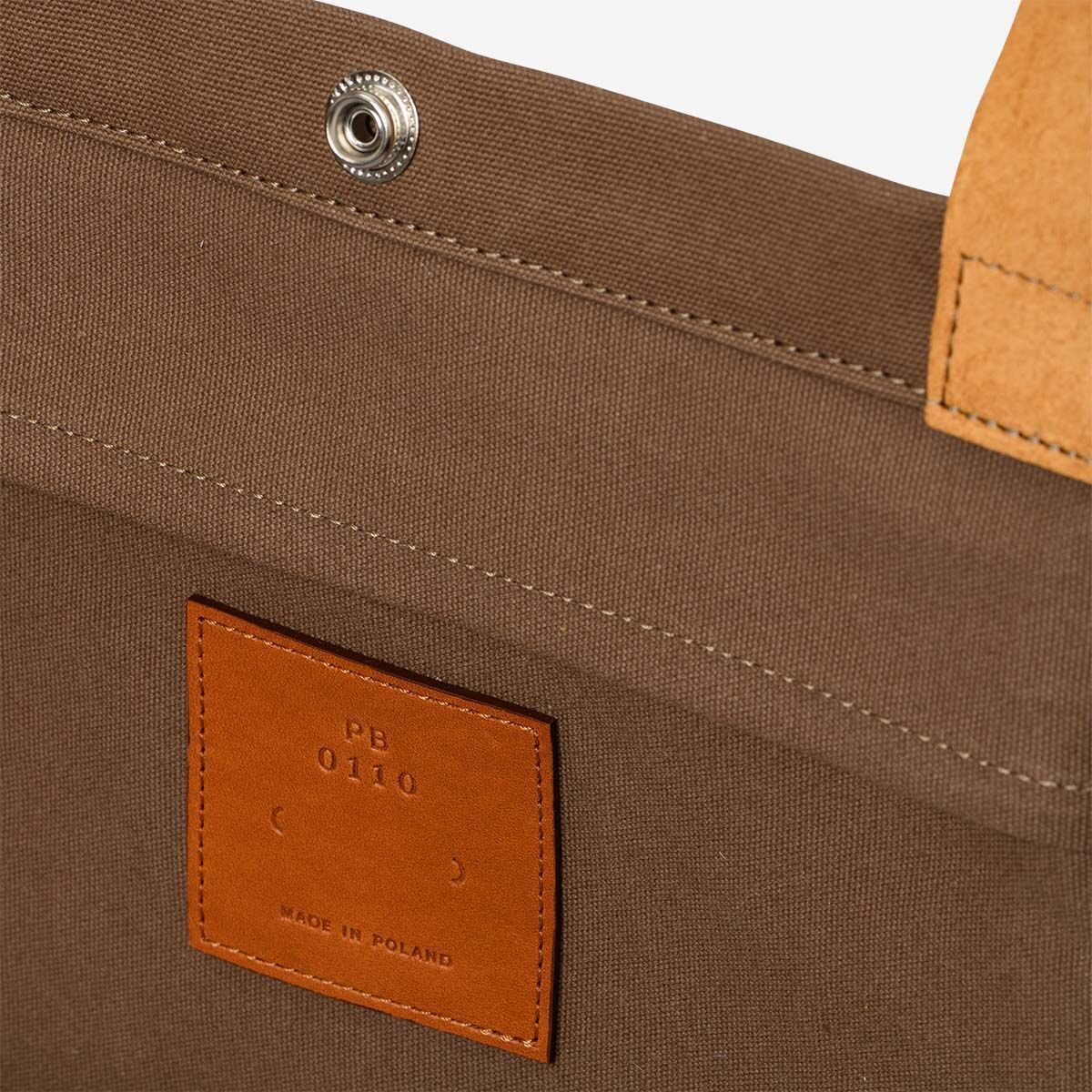 Eine detailreiche Aufnahme einer Jubiläums-Markttasche aus Stoff in Brauntönen und beigen Akzenten. Die Träger sind aus einem helleren Material gefertigt und ein dekorativer Druckknopf ist sichtbar. Ein Etikett auf der Innenseite zeigt 'PB 0110' und 'Made in Poland'.