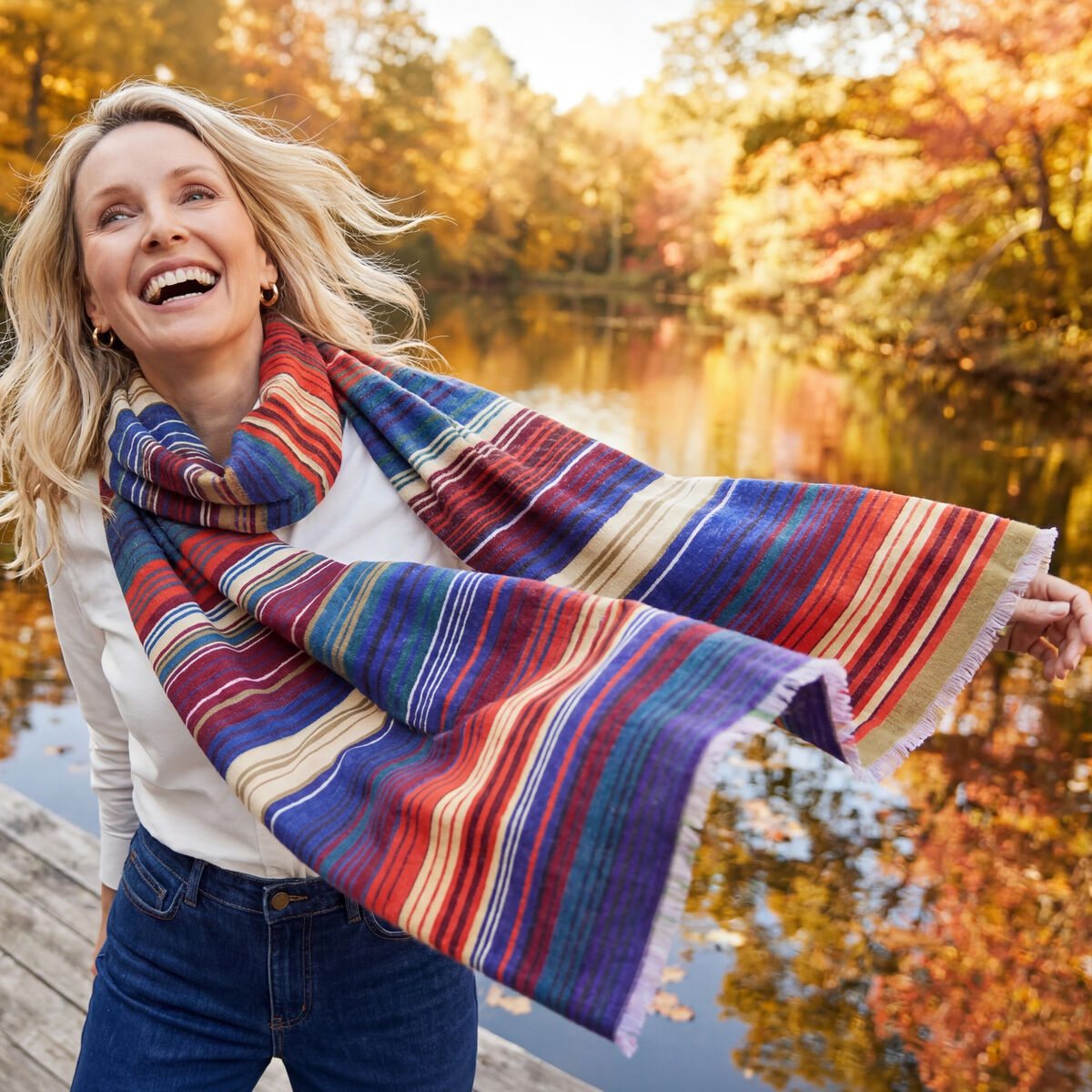Eine lachende Frau trägt den großformatigen Wollschal »Ariana« von Eagle Products locker um den Hals. Der weiche Schal aus Merino-Schurwolle zeigt breite, unregelmäßige Streifen in Blau, Rot, Violett, Türkis und Creme. Sie steht auf einem Holzsteg an einem stillen See, umgeben von goldenem Herbstlaub und Spiegelungen im Wasser.