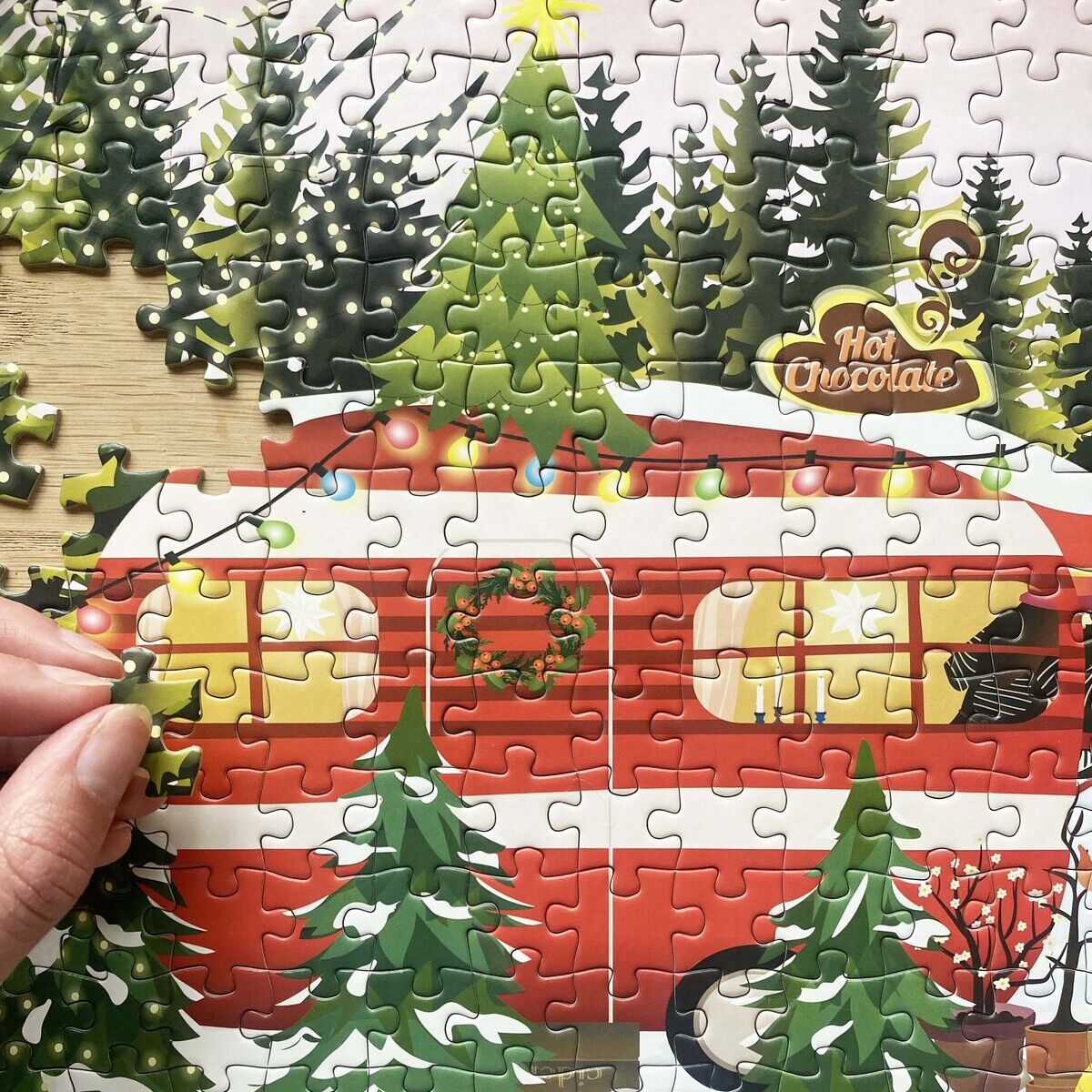 Detailreiches Puzzle mit 300 Teilen von VISSEVASSE, Motiv: roter Wohnwagen im verschneiten Winterwald, dekoriert mit Weihnachtslichtern, Tannenbäumen und Hot Chocolate Schild. Eine Hand setzt gerade ein Puzzleteil ein. Warme, festliche Atmosphäre.