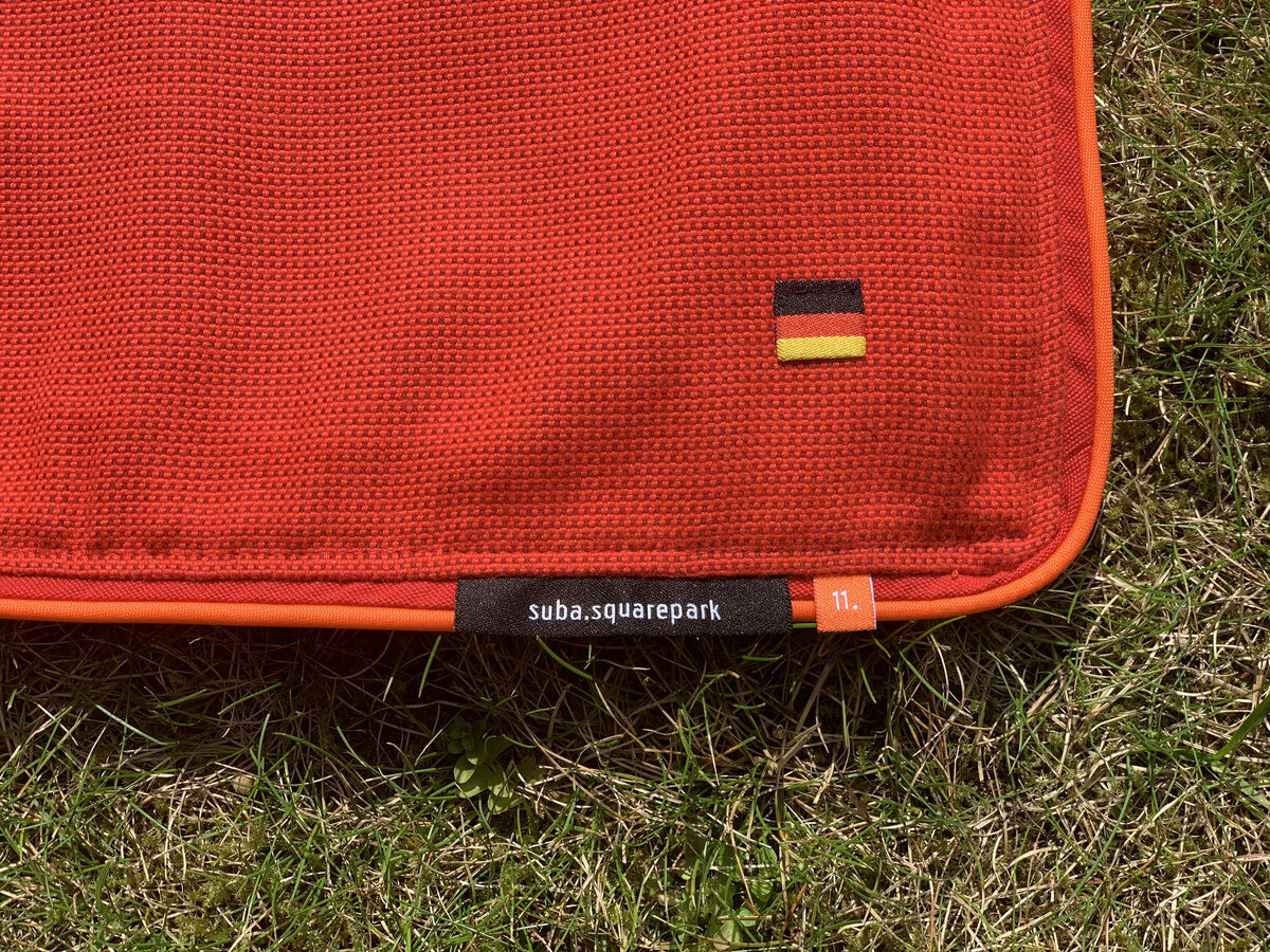 Nahaufnahme einer roten Picknickdecke namens „Squarepark 11“ auf einer grünen Wiese, mit einem kleinen deutschen Flaggenetikett und einem dunklen Stoffetikett.