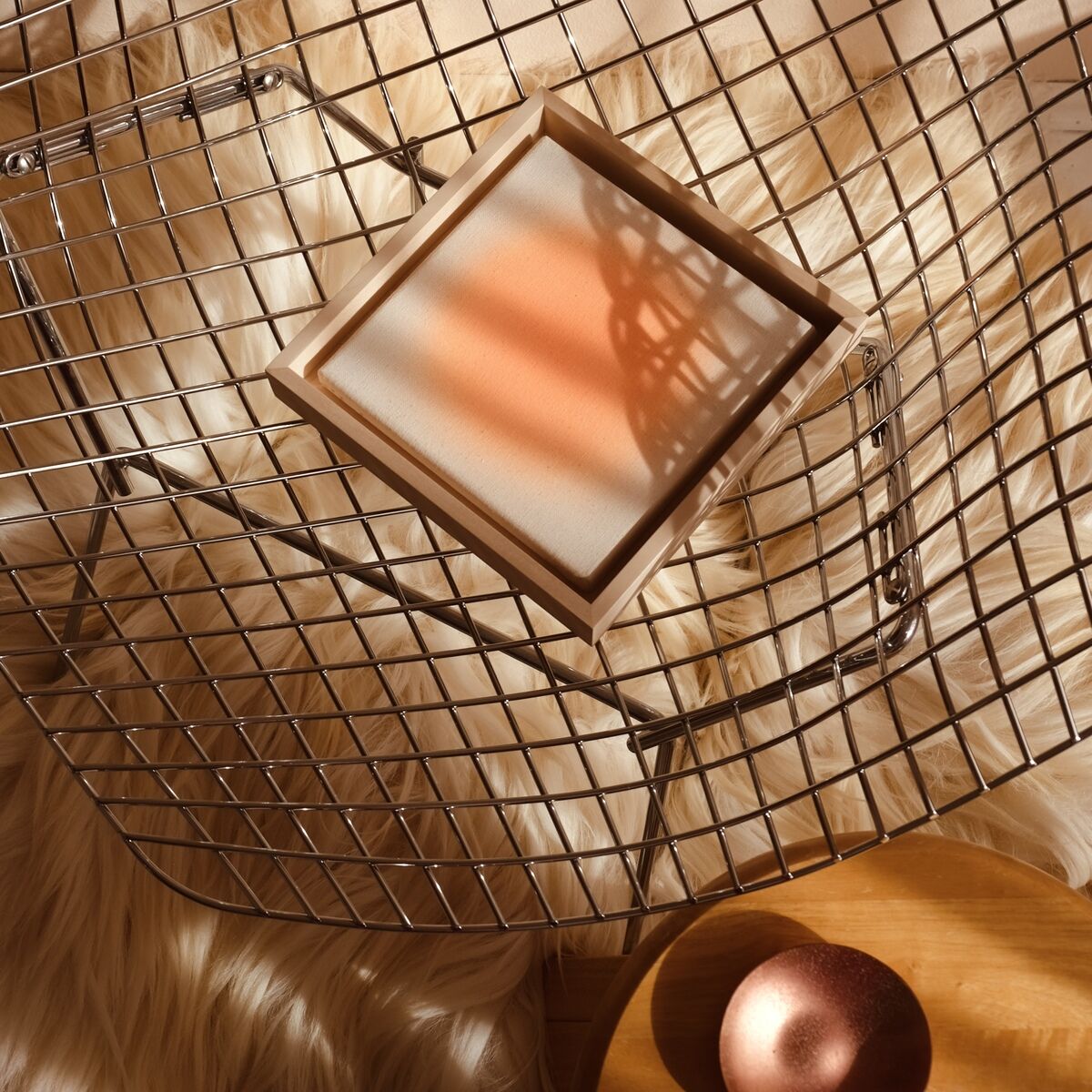 Quadratischer Kunstdruck in Sorbet-Orange-Tönen in einer hellen Umgebung, arrangiert auf einer modernen Metallablage mit flauschigem Untergrund und dekorativer Kugel, betont minimalistisches und stilvolles Wohndesign.