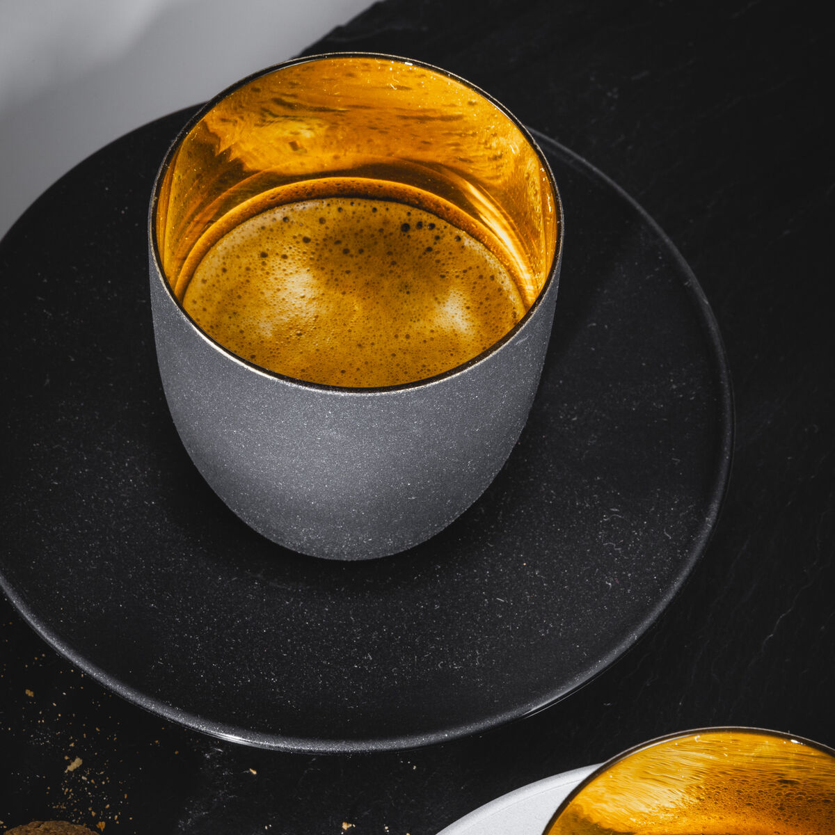 Zwei moderne Espressobecher auf schwarzen und weißen Untertassen, einer mit goldener Innenbeschichtung, gefüllt mit Espresso. Im Vordergrund befinden sich einige Keksstücke und Krümel. Das Bild ist stilvoll arrangiert und hebt die Eleganz der Kaffeetassen hervor.