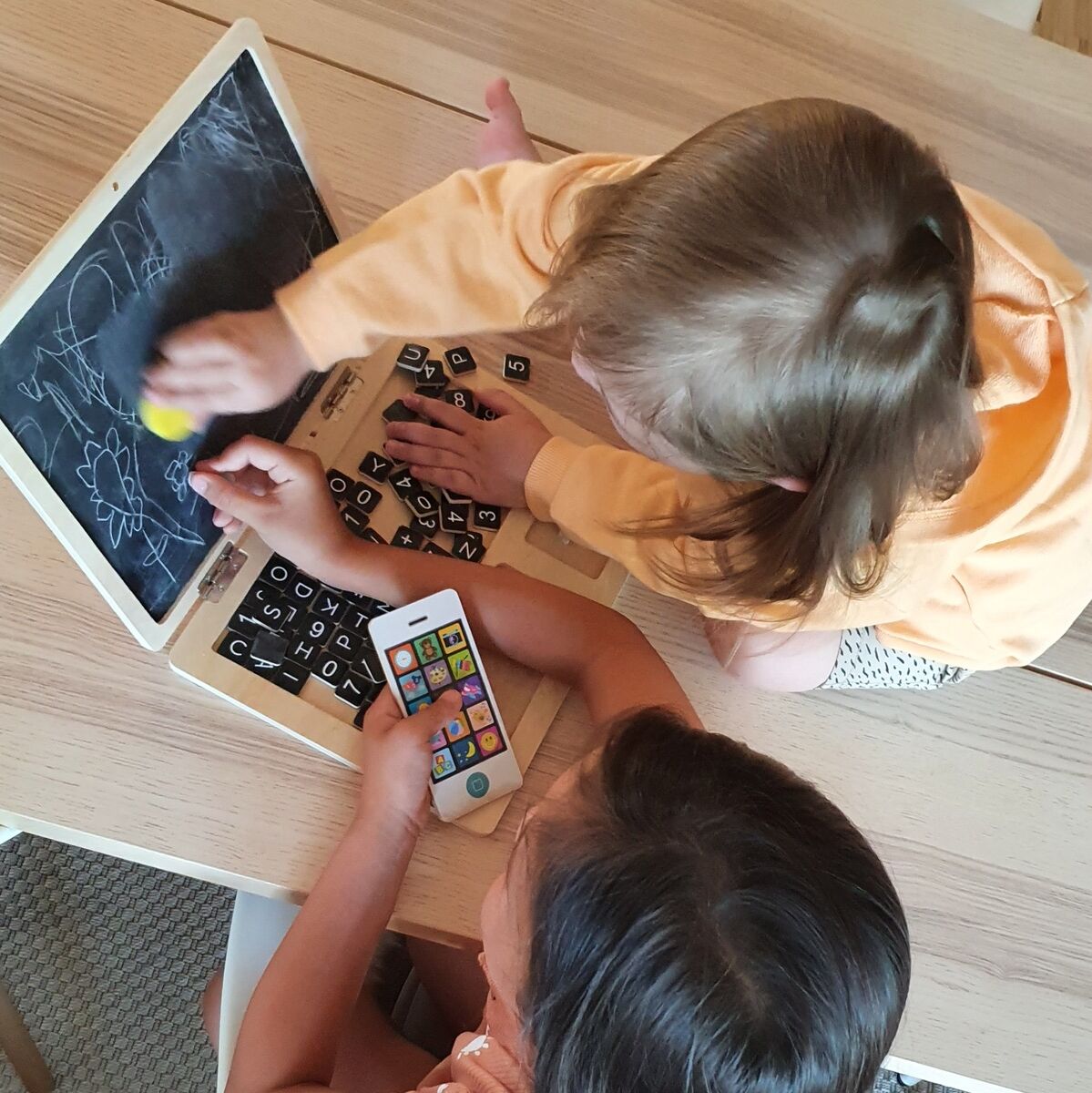 Zwei Kinder spielen an einem hölzernen Spielzeug-Laptop mit Magnettafel. Eines der Kinder malt auf der Tafel, während das andere Magnetbuchstaben sortiert.