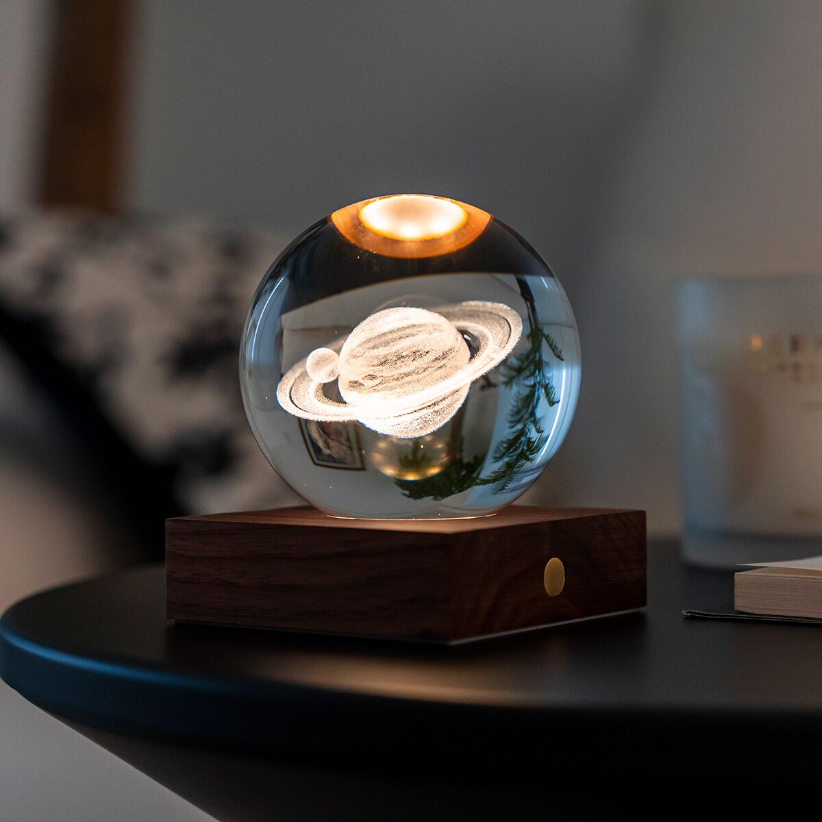 Auf einem Tisch steht eine moderne Lampe mit einer schwebenden Glaskugel, die von innen beleuchtet ist und die Saturnringe darstellt. Die Basis der Lampe besteht aus dunkelbraunem Holz mit einem goldfarbenen Knopf. Die Lampe strahlt warmes Licht aus und sorgt für eine gemütliche Atmosphäre in einem stilvoll eingerichteten Raum.