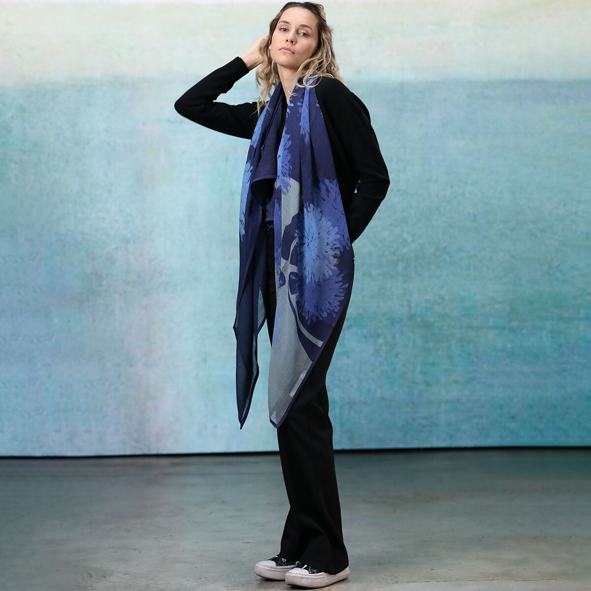 Eine Frau trägt einen großen, blauen Wollschal mit floralem Muster. Sie steht in einem minimalistischen Innenraum vor einer blau-grünen Wand und hat eine entspannte Pose. Der Schal wirkt weich und ist um Hals und Schultern gelegt. Das Outfit ist lässig und dunkel gehalten.