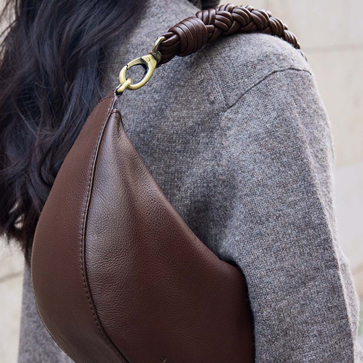 Eine Frau trägt eine elegante, dunkelbraune Schultertasche aus Leder mit geflochtenem Tragegurt und goldener Metallschnalle über einem grauen Strickpullover. Das Bild hebt den modernen und klassischen Stil der Tasche hervor.
