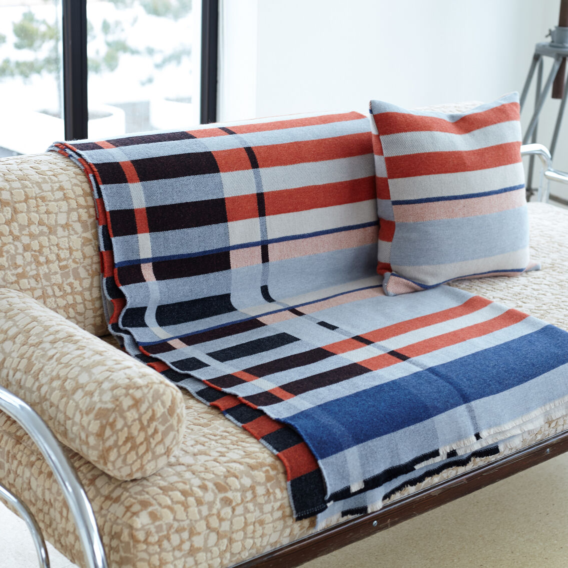 Eine gewebte Decke und ein passendes Kissen mit buntem Streifenmuster im Bauhaus-Stil liegen auf einem cremefarbenen Sofa mit metallischem Gestell vor großen Fenstern.