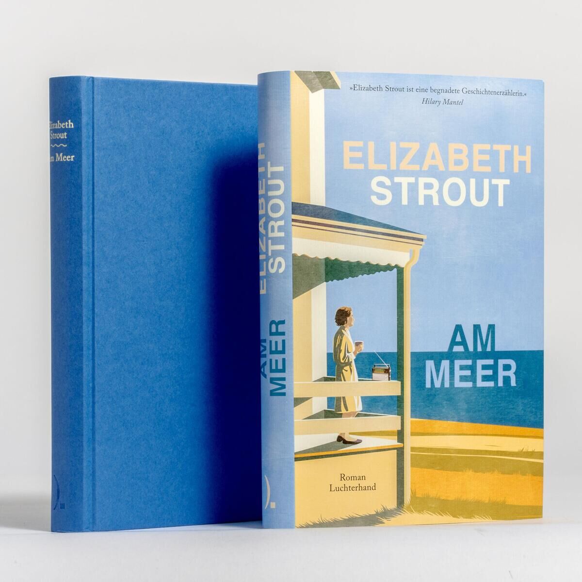 Das Bild zeigt das Buch 'Am Meer' von Elizabeth Strout. Das Cover des Buches zeigt ein Strandhaus mit einem Blick auf das Meer und einen Menschen, der auf die Ferne schaut. Das Buch hat einen blauen Hardcover-Einband.