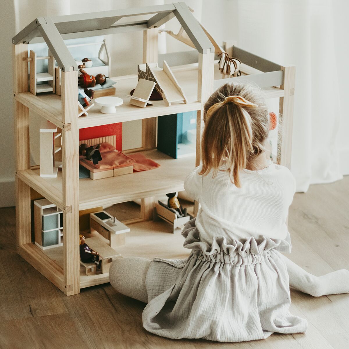 Ein Puppenhaus aus Holz mit mehreren Zimmern und modernen Miniaturmöbeln steht in einem hellen Raum. Ein Kind sitzt davor auf dem Boden und spielt konzentriert mit dem Spielzeughaus. Das Zimmer wirkt freundlich und einladend, die Farben sind hell und natürlich.