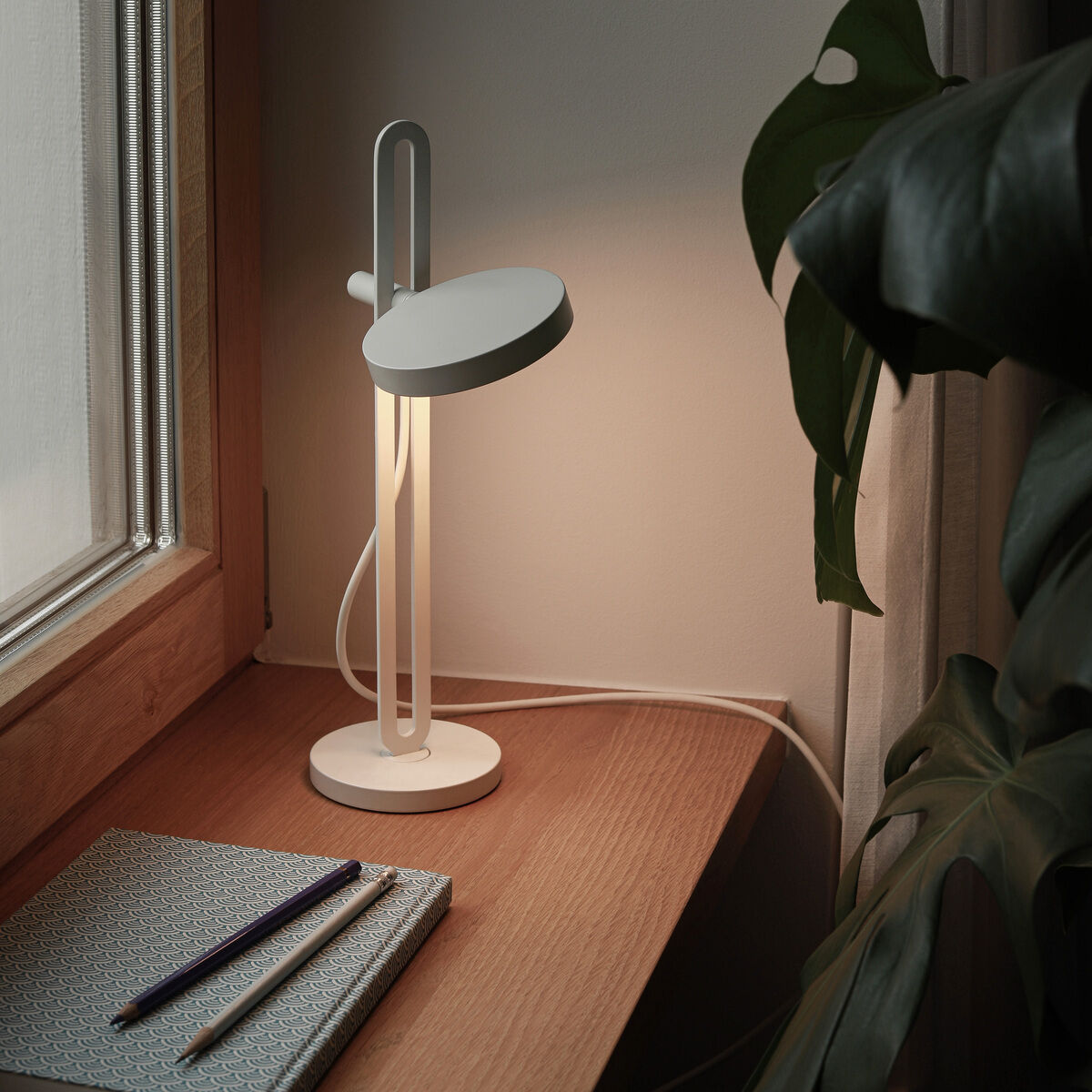 Ein modernes und minimalistisch gestaltetes Zimmer mit einer weißen Tischlampe namens »Echo«, die auf einem Holztisch neben einem Fenster steht. Ein Notizbuch und zwei Stifte liegen neben der Lampe. Eine große Pflanze mit grünen Blättern ist im Vordergrund sichtbar.