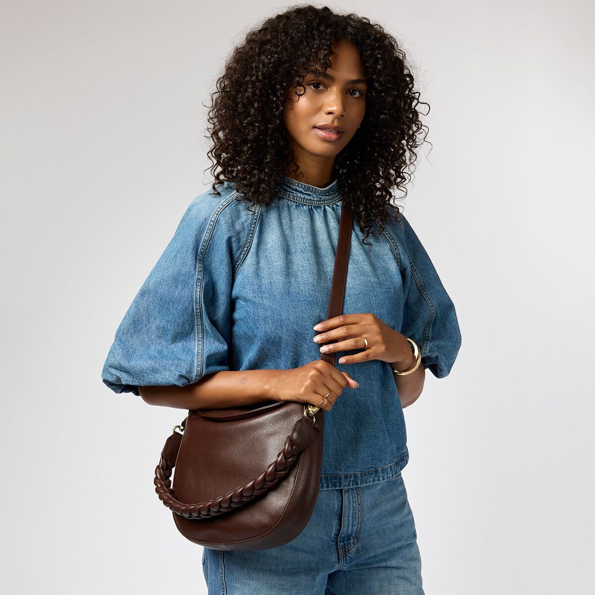 Eine Frau trägt eine dunkelbraune Umhängetasche aus Leder mit geflochtenem Riemen über der Schulter. Sie ist in einem lässigen Outfit mit Jeans und einem blauen Oberteil gekleidet und steht vor neutralem Hintergrund. Die Tasche ist schlicht, praktisch und modisch gestaltet.