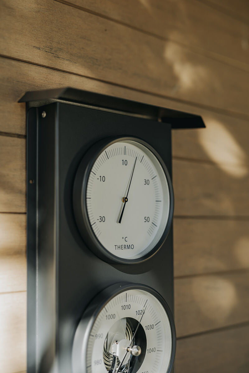 Nahaufnahme einer Außenwetterstation, durch die Holzwand montiert. Sie verfügt über zwei analoge Messgeräte zur Anzeige der Temperatur, mit klarer Skala und Zeigern. Schattierungen der Sonne sind auf der Holzverkleidung sichtbar.