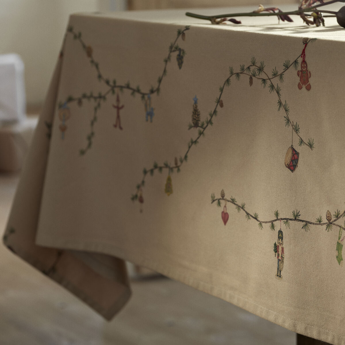 Beigefarbene Tischdecke mit gestickten grünen Zweigen und bunten Weihnachtsmotiven, dekorativ auf einem Esstisch arrangiert, sanft beleuchtet, geeignet für festliche Anlässe.