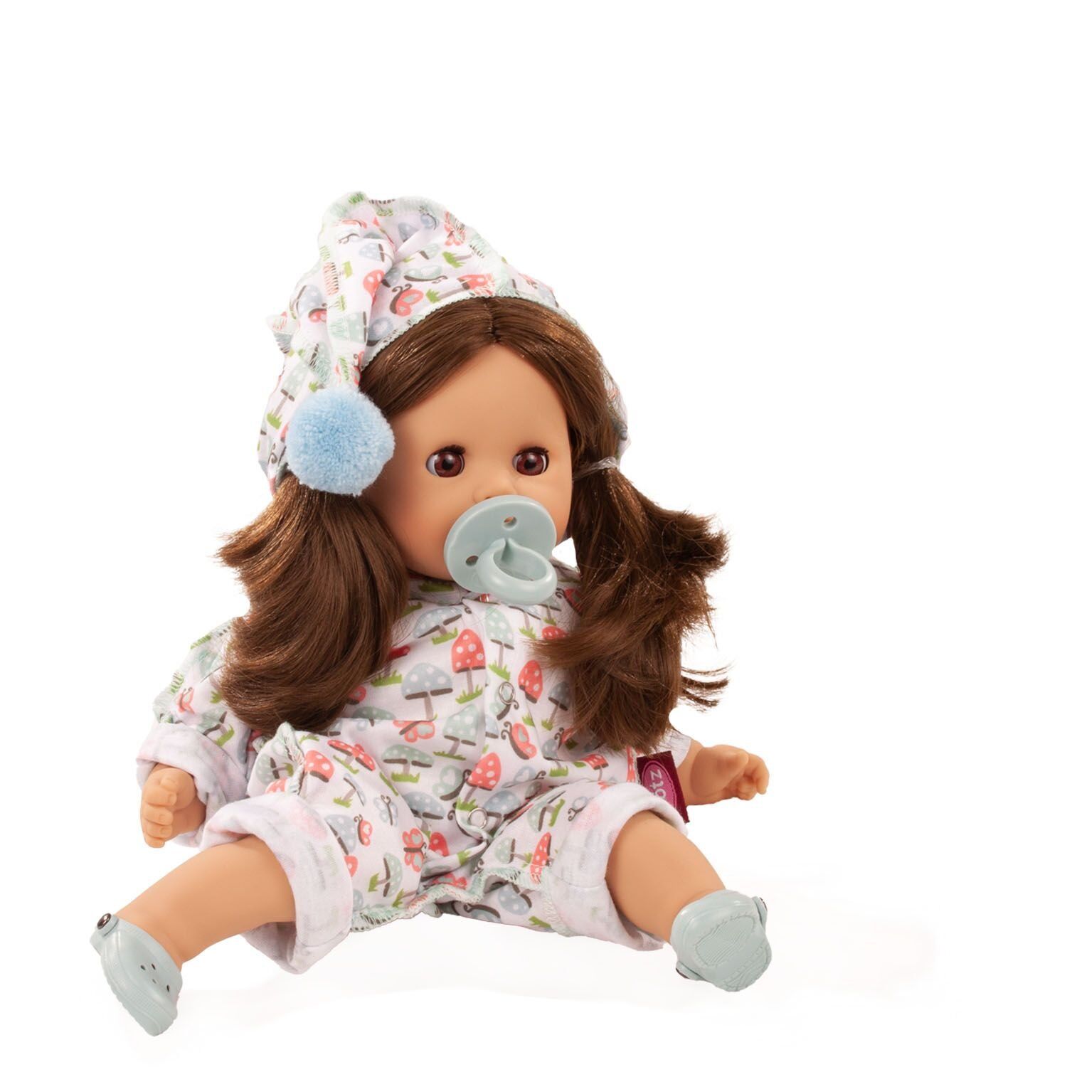 Eine weiche Badepuppe, angezogen mit einem gemusterten Schlafanzug mit Pilzen und einer passenden Mütze mit Bommel, hält einen Schnuller im Mund und sitzt aufrecht. Die Puppe eignet sich als Spielzeug für kleine Kinder und ist waschbar.