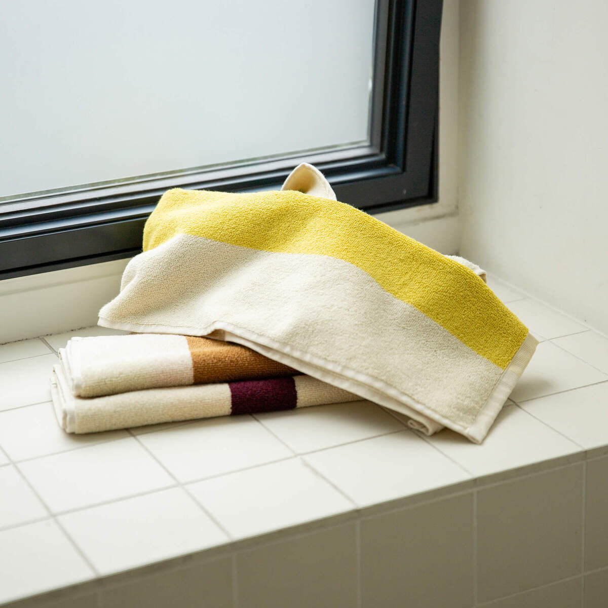 Ein Set von Gästehandtüchern in verschiedenen Farben, drapiert auf einer weißen Kachelfläche neben einem Fenster. Die Handtücher sind überwiegend weiß mit Streifen in Gelb, Braun, und Bordeauxfarbtönen.