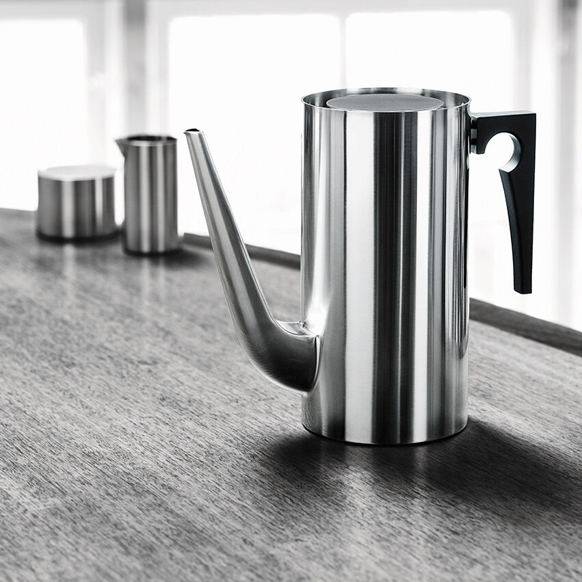 Eine stilvolle Kaffeekanne im modernen Design von Arne Jacobsen, hauptsächlich aus Edelstahl mit einem schwarzen Griff. Die Kanne steht auf einer Holztischplatte mit weiteren Edelstahlobjekten im Hintergrund. Das Design ist minimalistisch und zeitlos.