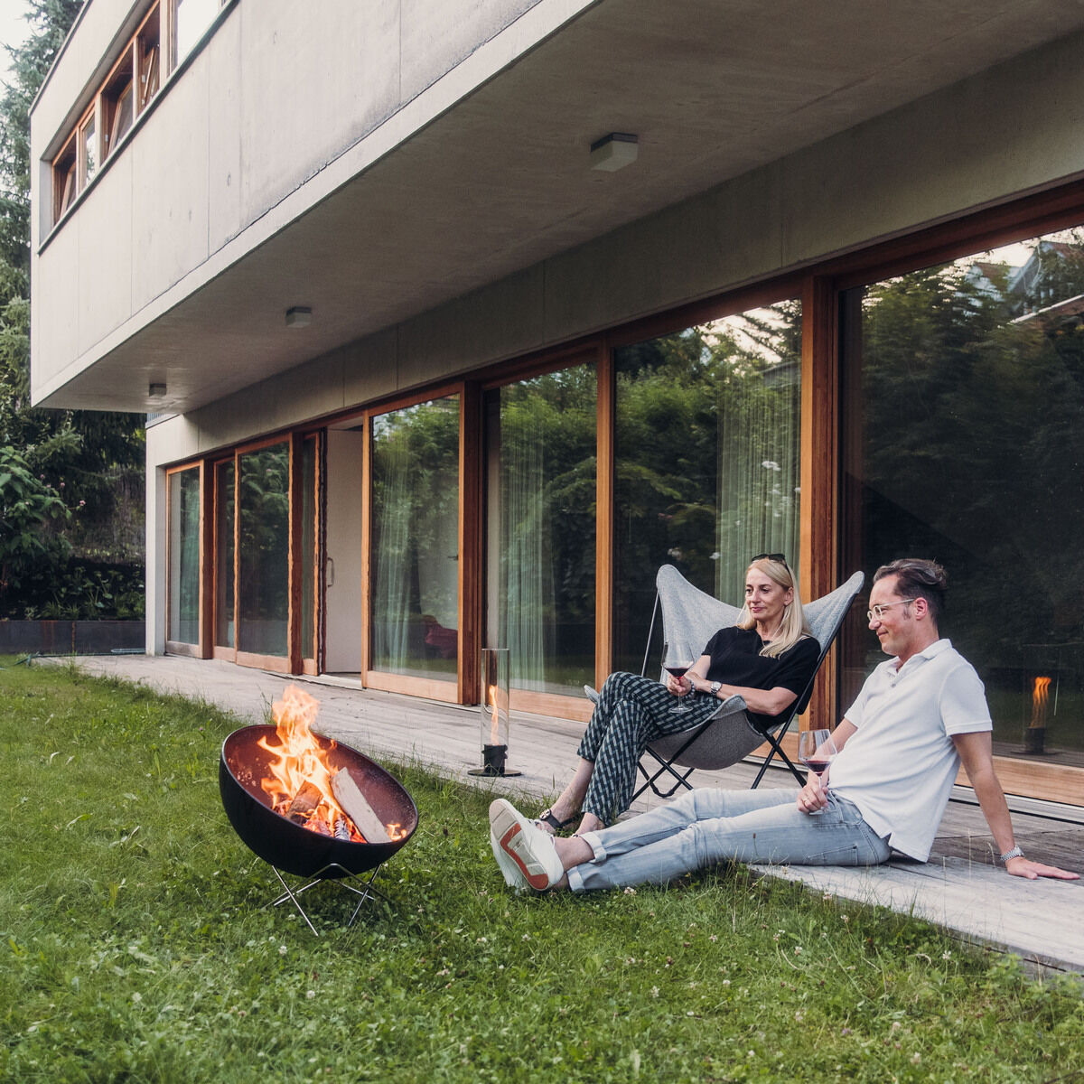 Ein modernes Haus mit großen Fenstern und Holzbalkon, davor eine Grasfläche mit einer brennenden Feuerschale »BOWL 57«. Zwei Personen sitzen entspannt auf Klappstühlen neben der Feuerschale.