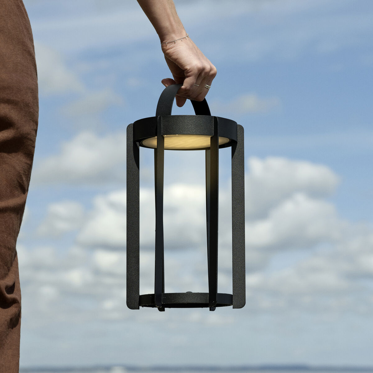 Eine Person hält eine moderne LED-Laterne »Firefly« mit schmalem Design, die sich gegen den Hintergrund eines klaren, blauen Himmels mit vereinzelten Wolken abhebt. Die Laterne hat eine schwarze Struktur und strahlt warmes, gelbes Licht aus.