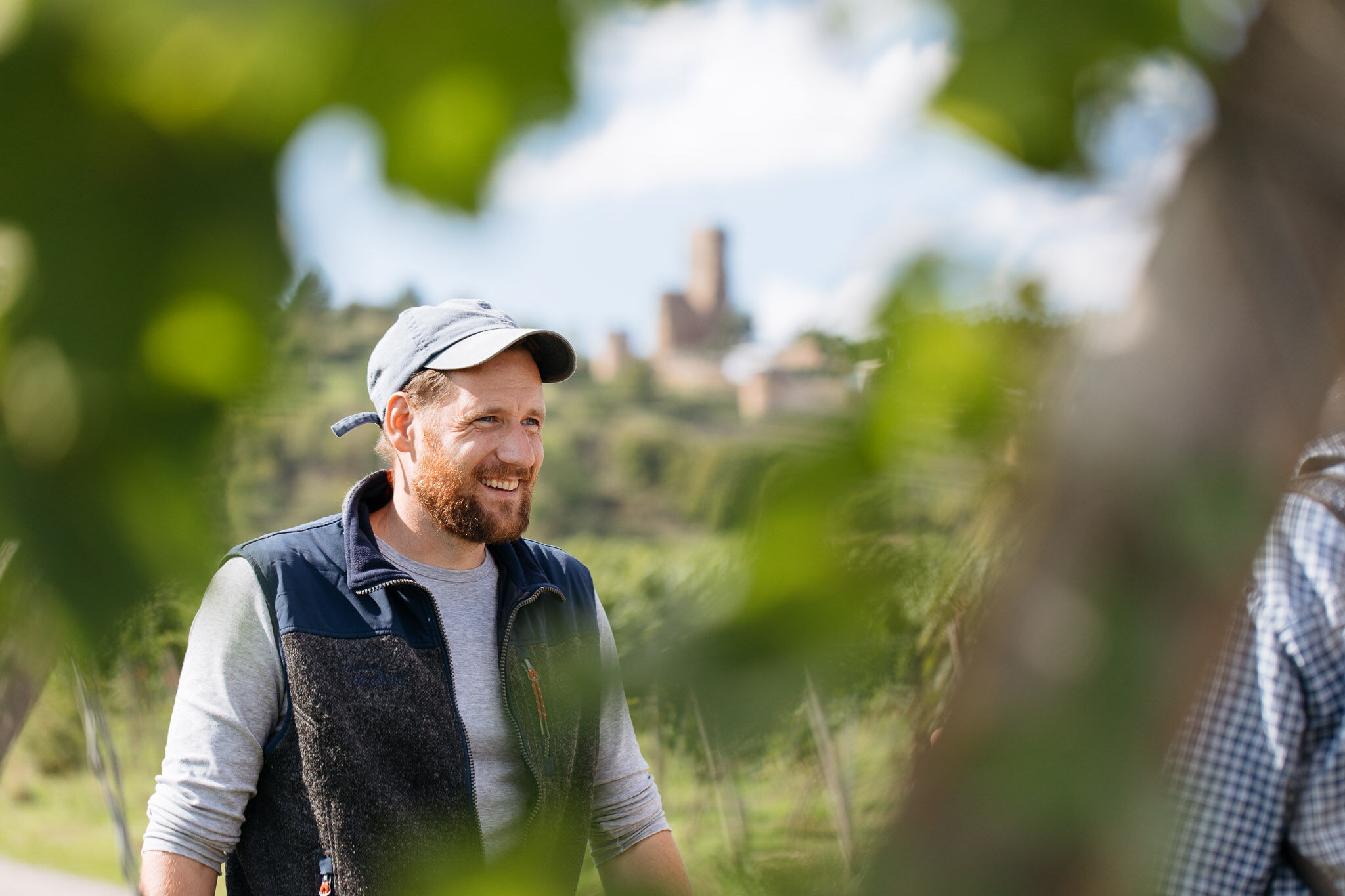 Person mit grauer Kappe und Weste steht in einem Weinberg, im Hintergrund eine Burg auf einem Hügel