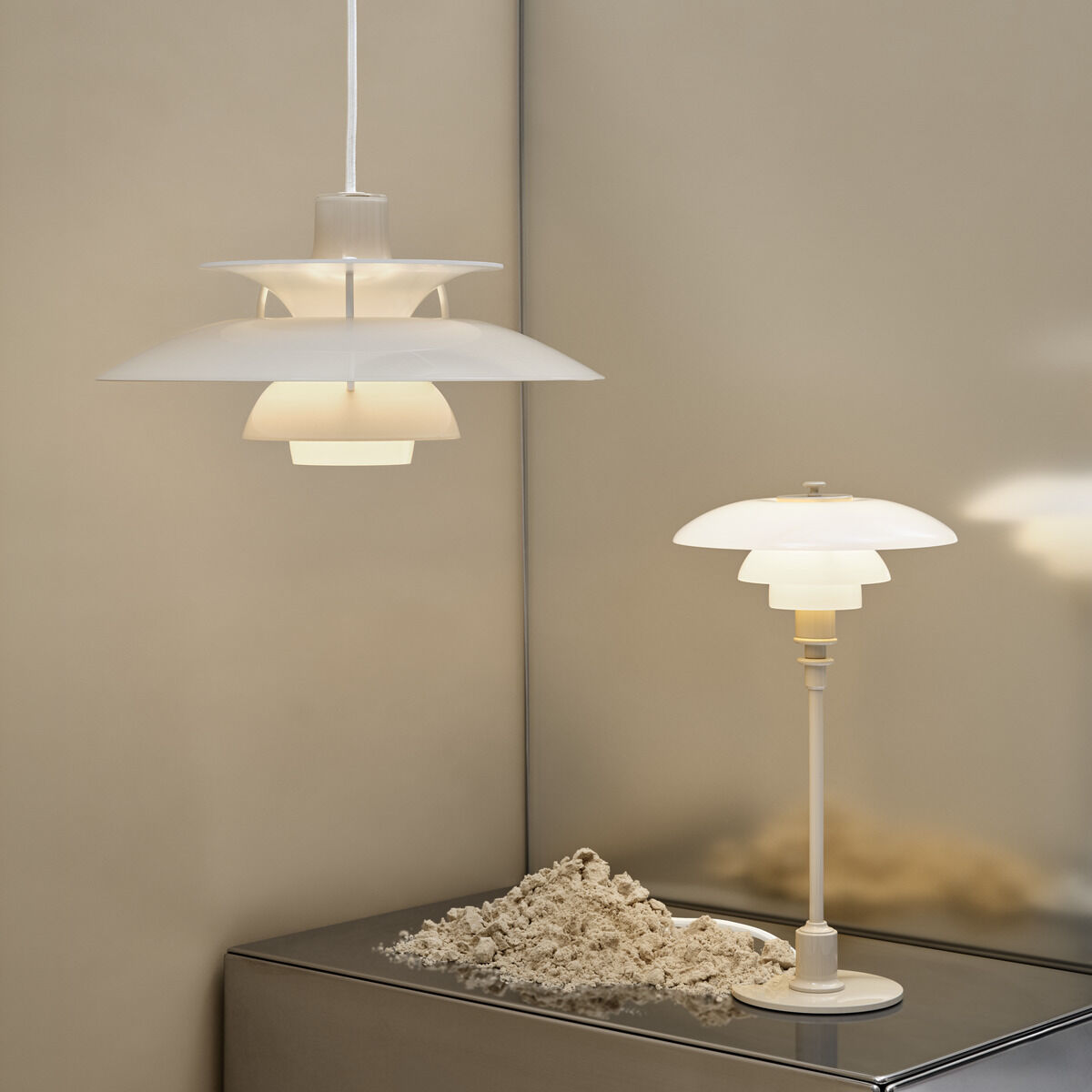 Die Hängeleuchte »PH 5 320« in opal beige hängt vor einer beigen Wand, daneben steht eine farblich passende Tischleuchte auf einer sandbedeckten, metallischen Oberfläche. Beide Lampen haben ein ikonisches mehrschichtiges Design und strahlen warmes Licht aus.