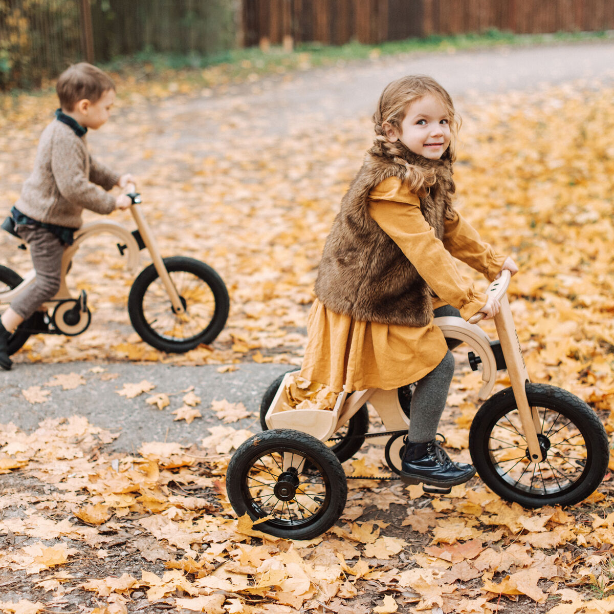 Zwei kleine Kinder fahren draußen im Herbst auf einem Balance Bike und einem Dreirad mit Erweiterung über einen mit gelben Blättern bedeckten Weg. Die Szene zeigt fröhliches Spielen, Bewegung und gemeinsames Entdecken im Freien.