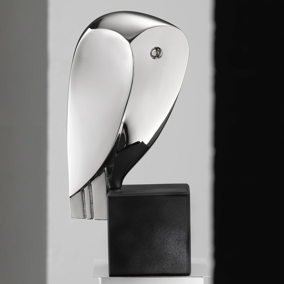Abgebildet ist eine elegante, moderne Skulptur eines stilisierten Vogels der Künstlerin Cordula Dobes in metallic-silber auf einem schwarzen, quaderförmigen Sockel. Die Oberfläche glänzt spiegelnd. Die Skulptur steht vor einem minimalistischen, hell-dunkel geteilten Hintergrund.