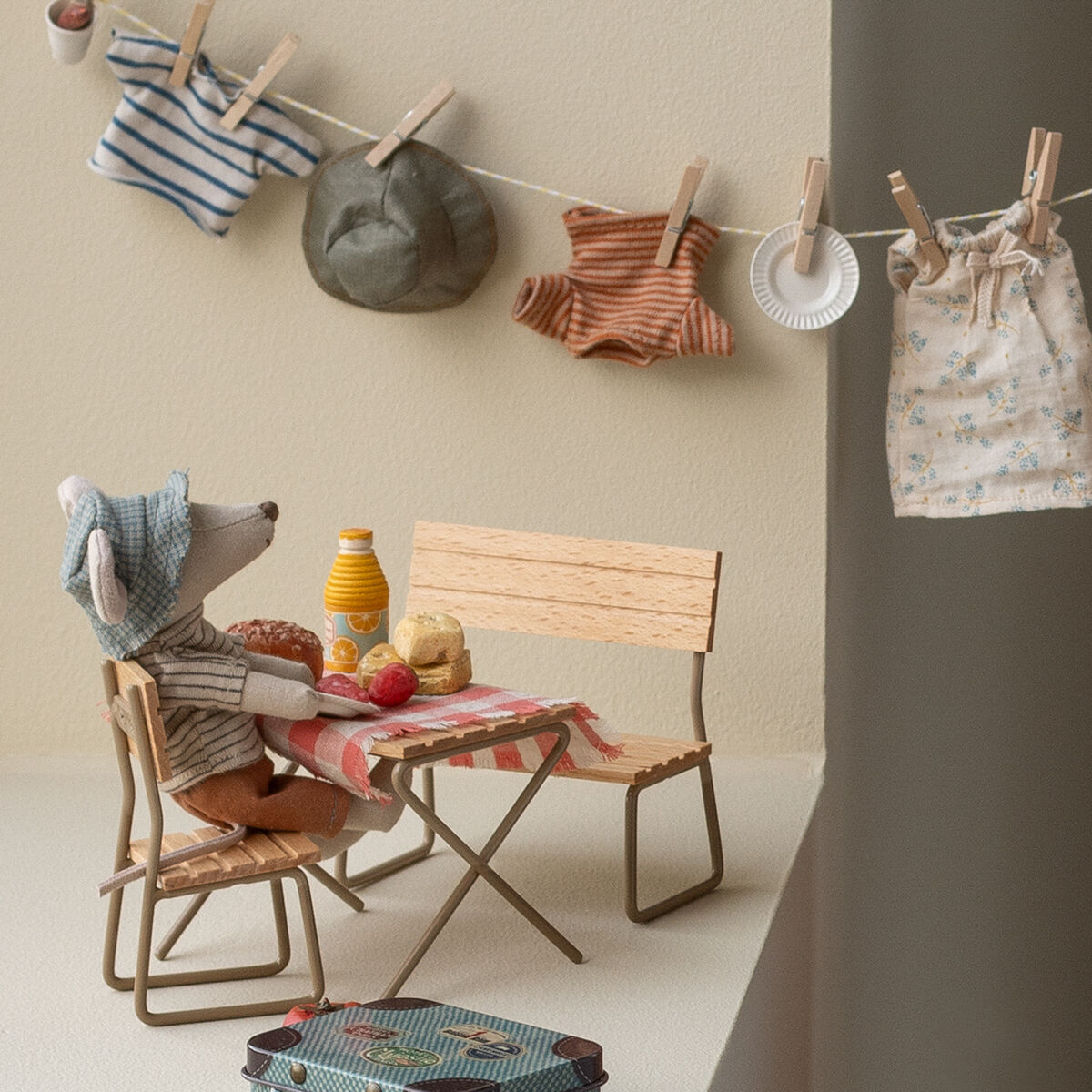 Auf dem Bild sitzt eine kleine Stoffmaus mit Hut und Rucksack an einem Miniatur-Holztisch, der mit einem karierten Tischtuch, Brötchen, Getränk und Obst gedeckt ist. Im Hintergrund hängen Kinderkleidung und ein Hut auf einer Wäscheleine. Die Szene wirkt liebevoll gestaltet und detailreich, ideal für Kinder oder Sammler von Maileg-Figuren.