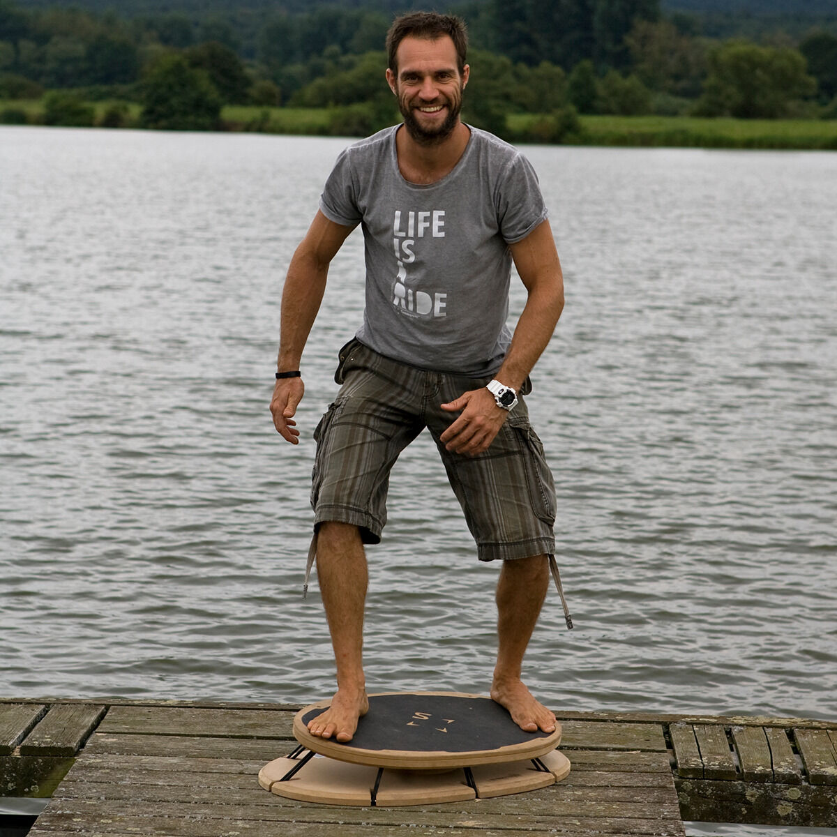 Ein Mann steht auf einem Sensoboard »essential« auf einem hölzernen Steg vor einem See. Er trägt ein graues T-Shirt mit der Aufschrift 'Life is Wide' und kurze graue Hosen. Im Hintergrund sind grüne Bäume und Berge zu sehen.