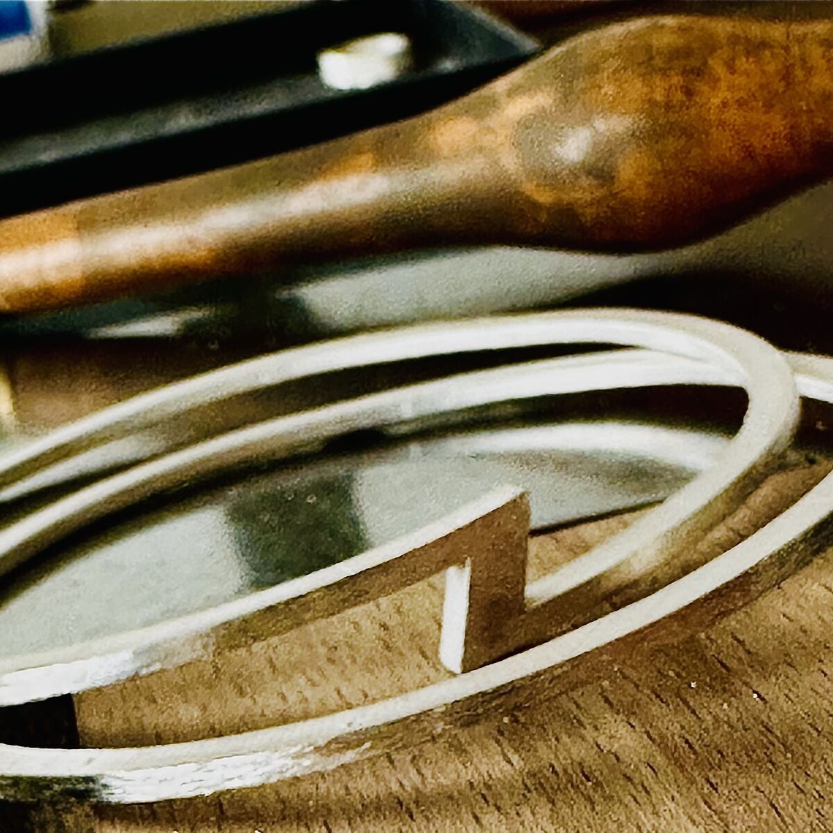 Detaillierte Nahaufnahme von mehreren silberfarbenen Armreifen mit markantem Z-förmigem Designelement auf einer Holzunterlage. Im Hintergrund Werkzeuggriff, Fokus auf die handgefertigte, exklusive ZEIT Sonderedition von HML Berlin.