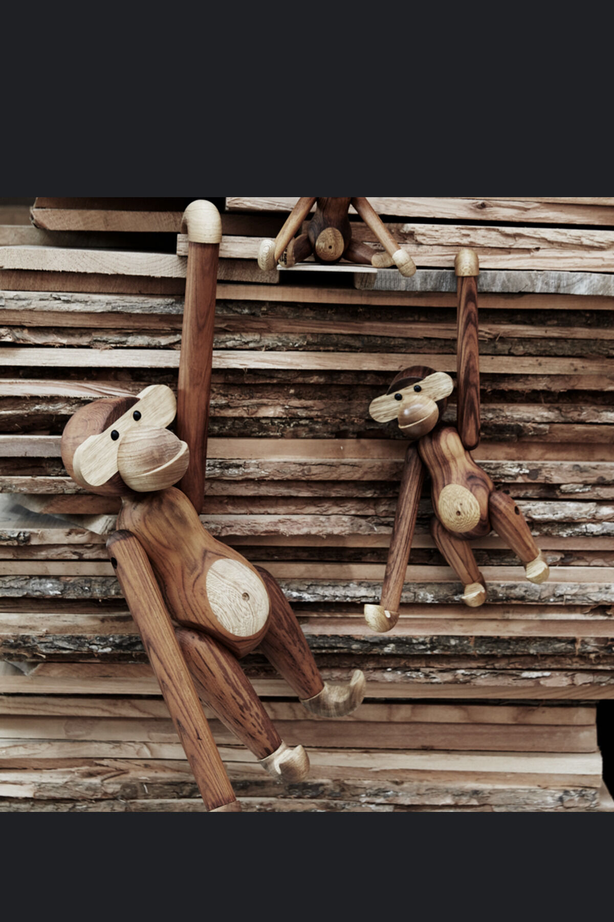 Drei Holzfiguren in Form von Affen, entworfen von Kay Bojesen, hängen und sitzen auf einem Stapel von Holzscheiten. Die Affen haben bewegliche Gliedmaßen und bestehen aus verschiedenen Holztönen.