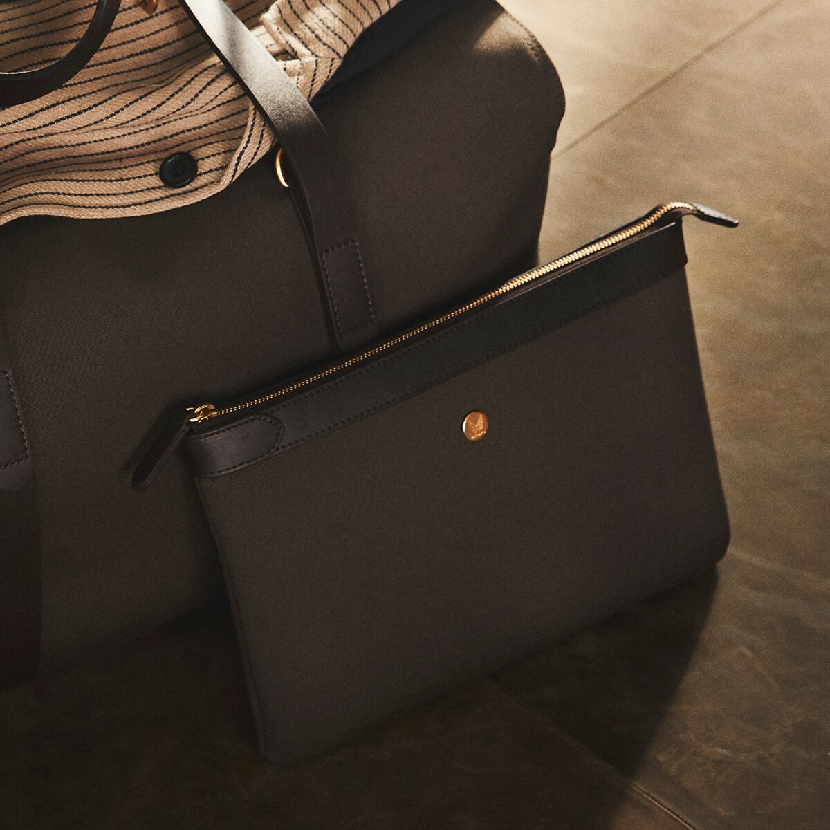 Große olivgrüne Canvas-Tasche mit dunkelbraunen Lederdetails und goldener Logoprägung. Im Vordergrund liegt die M/S Pouch Large von MISMO, elegant inszeniert auf dunklem Boden. Stabiles, wasserabweisendes Material und hochwertige Verarbeitung wirken zeitlos und luxuriös.