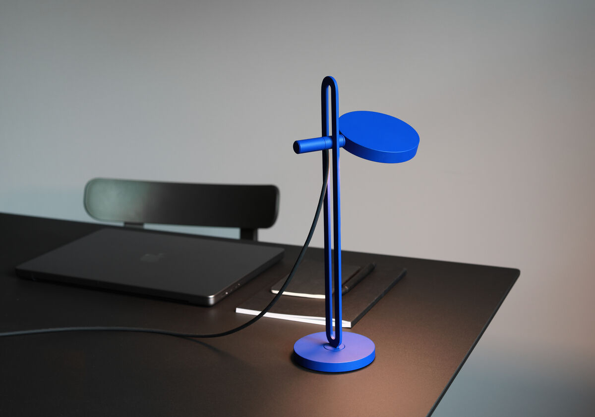 Ein modern gestaltete, blaue Tischlampe »Echo« steht auf einem schwarzen Tisch neben einem geschlossenen Laptop. Die Lampe hat ein minimalistisches Design mit einem runden Fuß und einem schlanken, gebogenen Arm. Das Ambiente ist schlicht, mit einem Hauch von Eleganz.