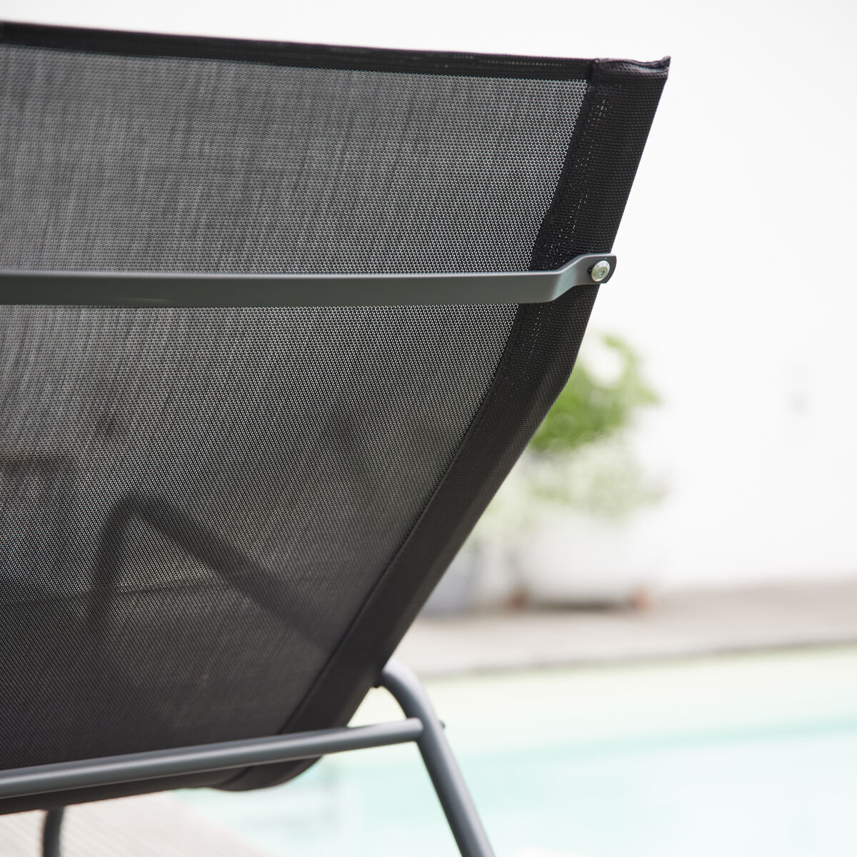 Seitliche Nahaufnahme einer modernen Swingliege »Aliano« aus schwarzem Netzstoff und grauem Metallrahmen, auf einer Holzterrasse am Pool. Im Hintergrund unscharf ein Paar Schuhe und ein grüner Blumentopf. Die Szene vermittelt sommerliche Entspannung im Außenbereich.